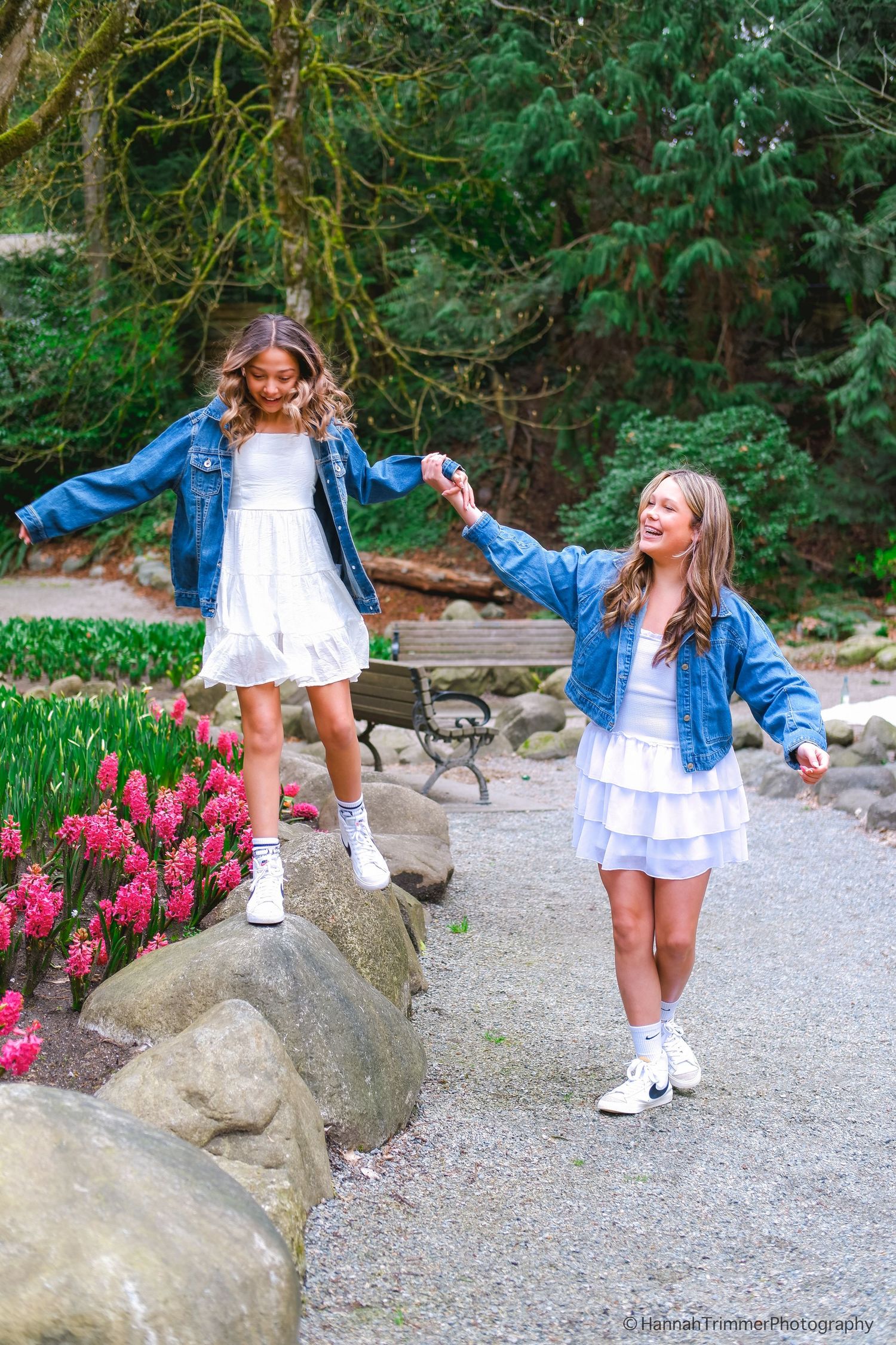 Two girls in white dresses and denim jackets laugh and play among pink flowers in a park.
