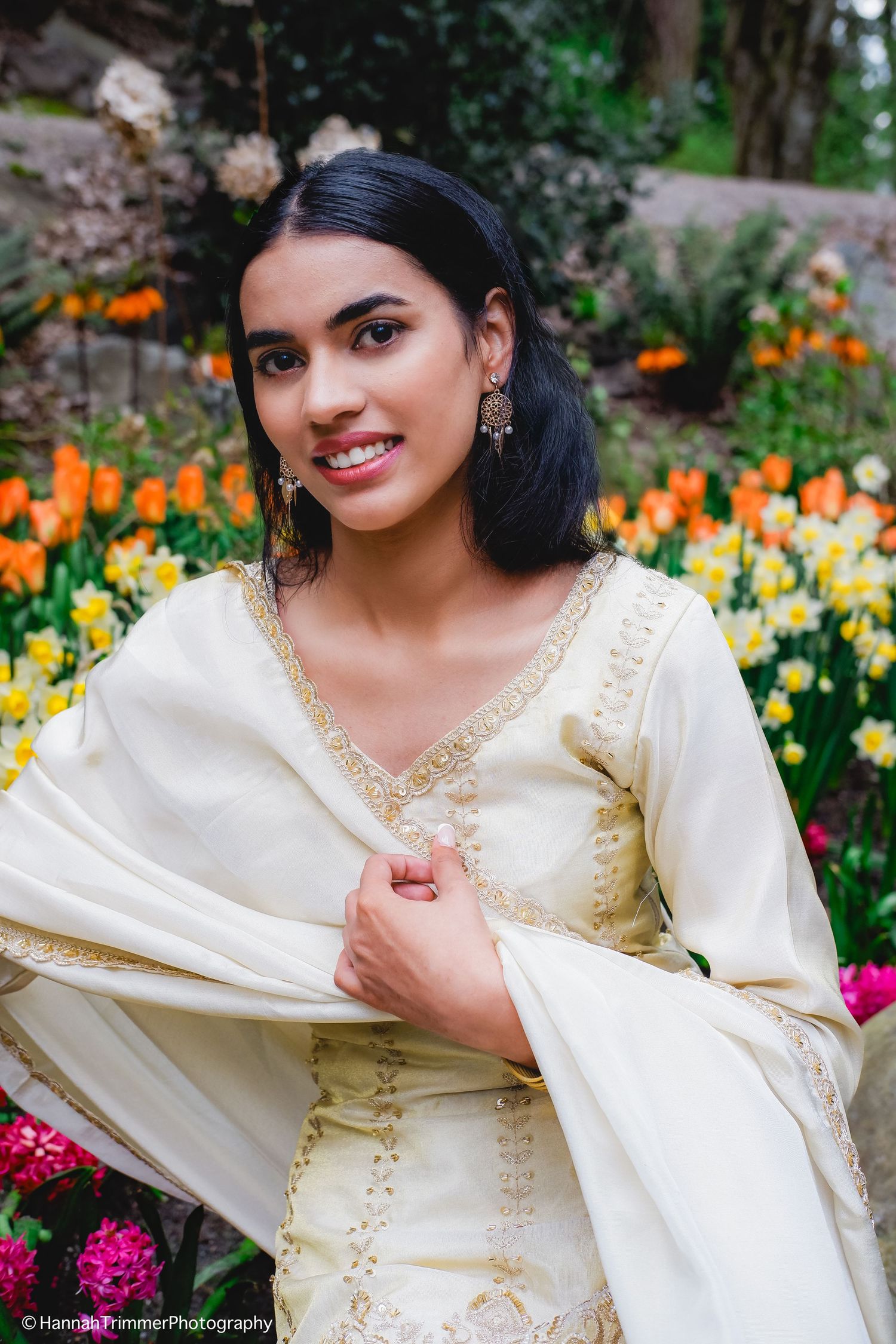 Young woman in cream embroidered salwar kameez smiles among vibrant tulips and daffodils.