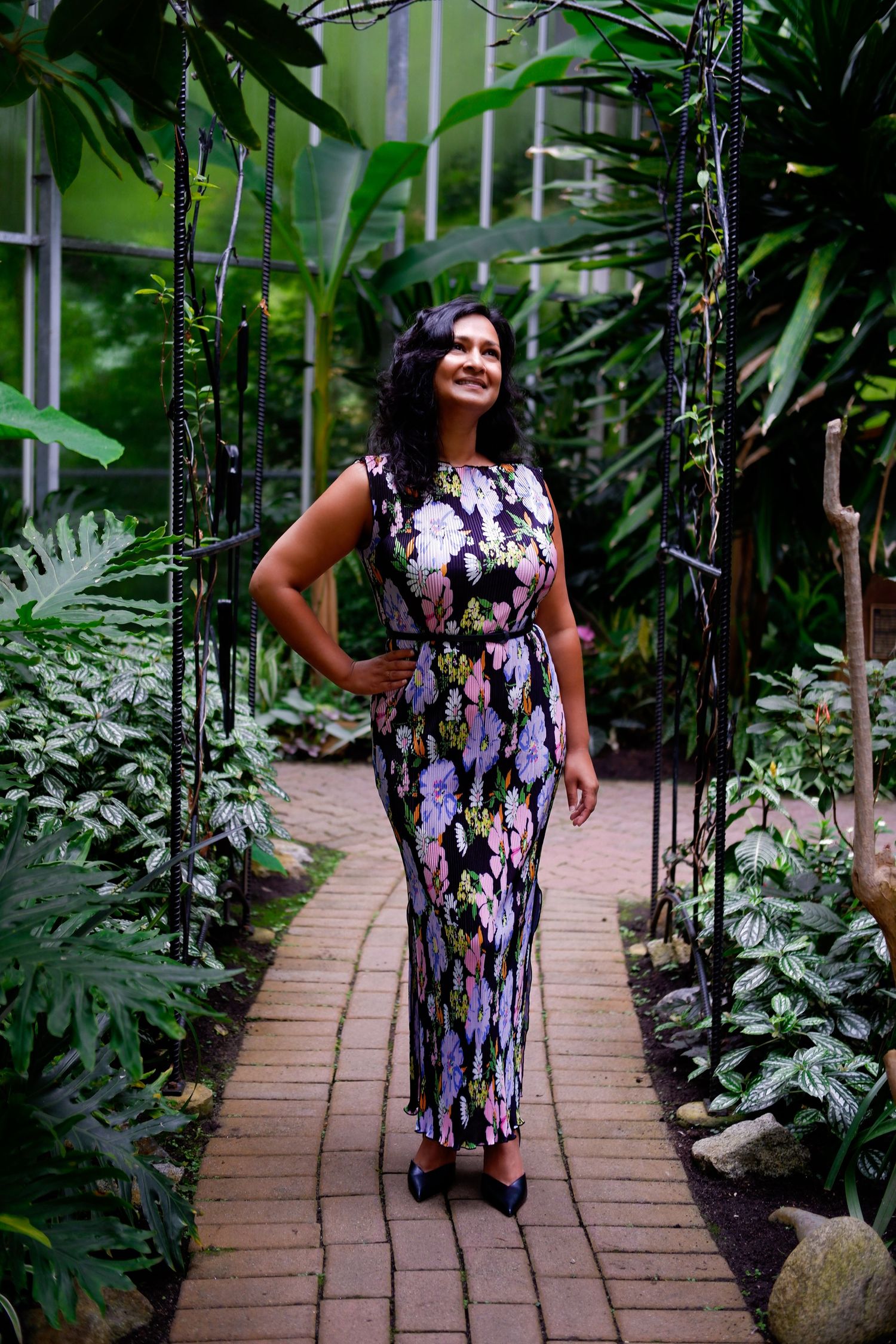 Woman in floral maxi dress standing in a tropical greenhouse archway.