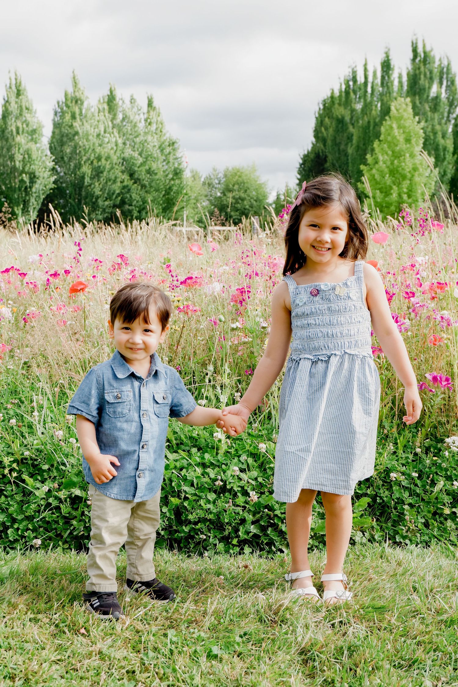 Two young siblings hold hands smiling in front of a colorful pink wildflower garden on a cloudy day.