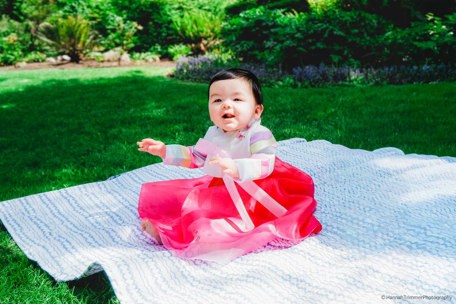 Adorable baby girl in pink Korean hanbok sits smiling on white blanket in green garden.