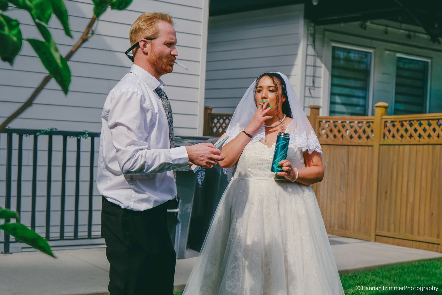 Red-haired groom hands drink to laughing bride in white wedding dress and veil outside a house.