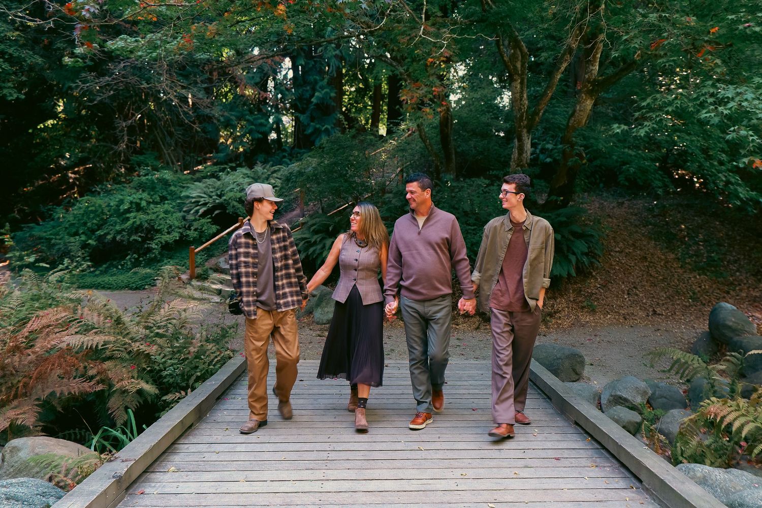 Four people walk together on a wooden bridge surrounded by lush green trees in a forest setting.