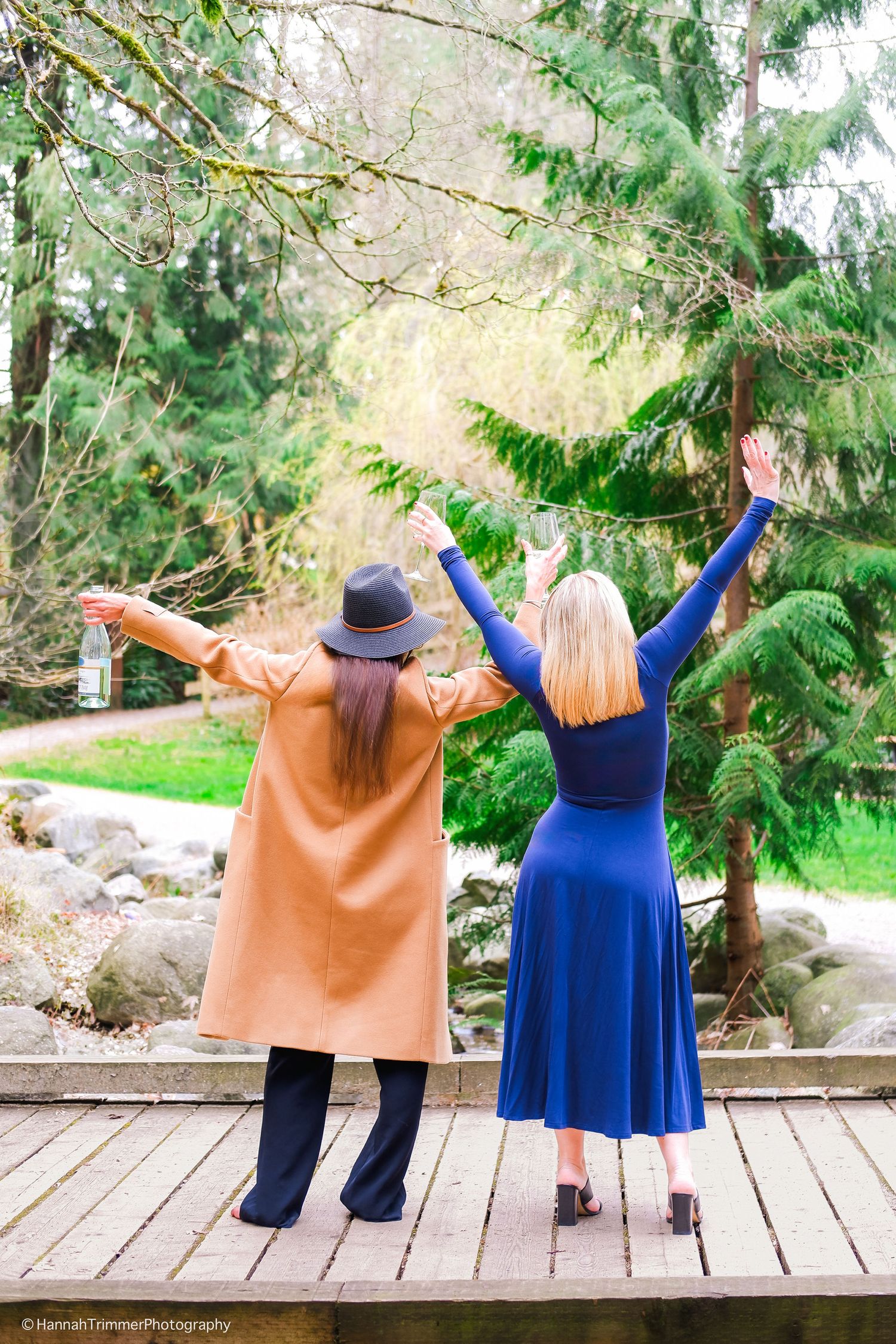 Two women with arms raised stand on a wooden deck surrounded by lush green trees in a garden.