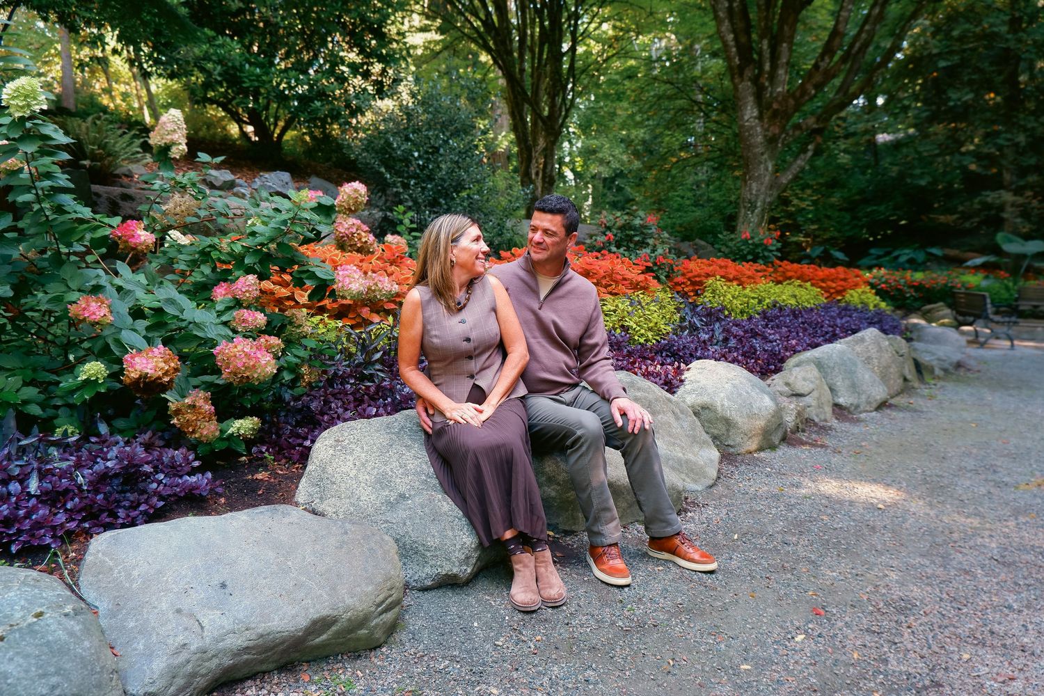 Couple sitting on rocks surrounded by vibrant colorful flower gardens in a lush green park setting.