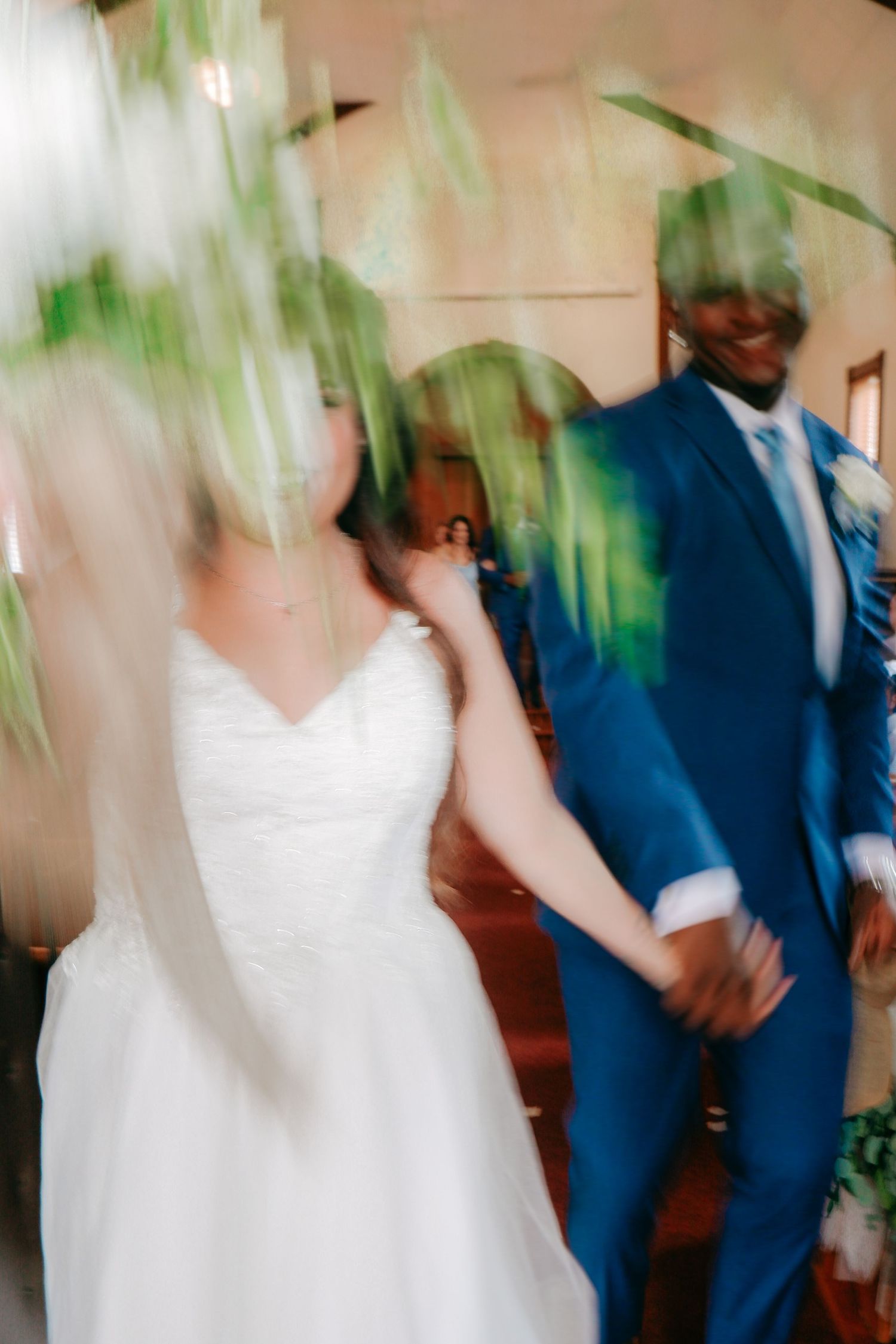 Blurred motion shot of interracial wedding couple holding hands, bride in white gown, groom in blue suit, walking down aisle.