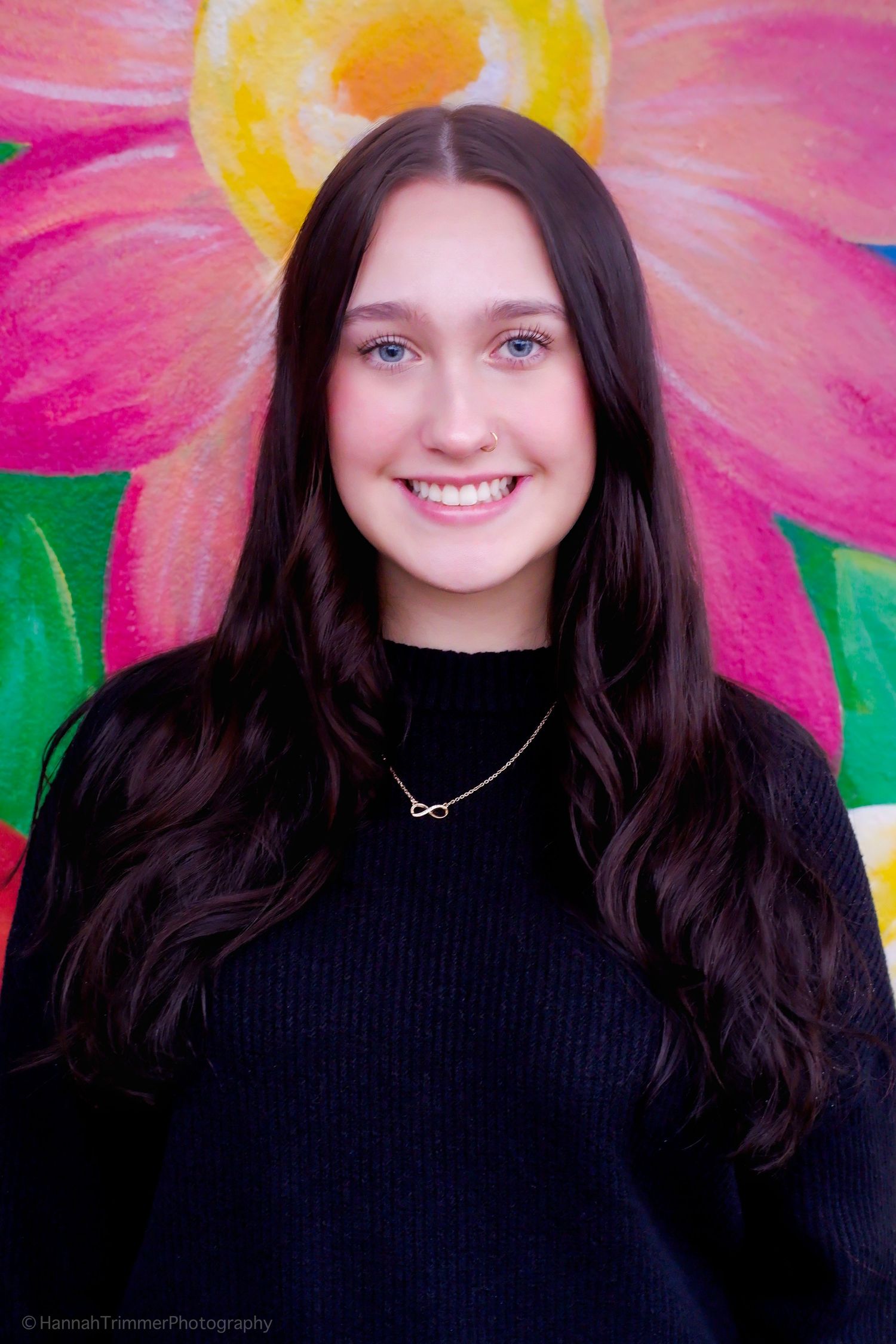 Smiling teenage girl with blue eyes and dark hair posing before a vibrant painted flower mural.