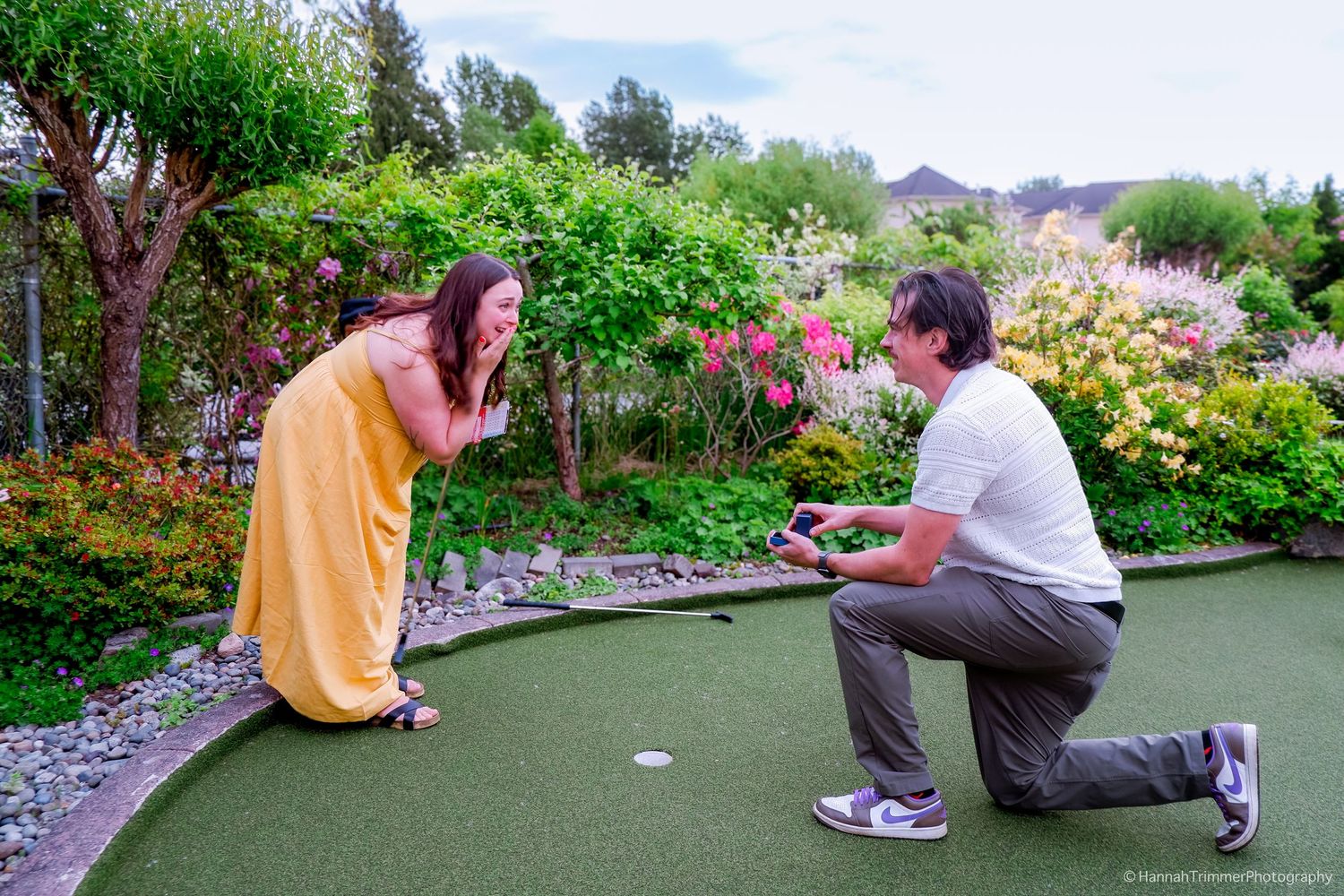 Man on one knee proposing to surprised woman in yellow dress on a mini golf course.