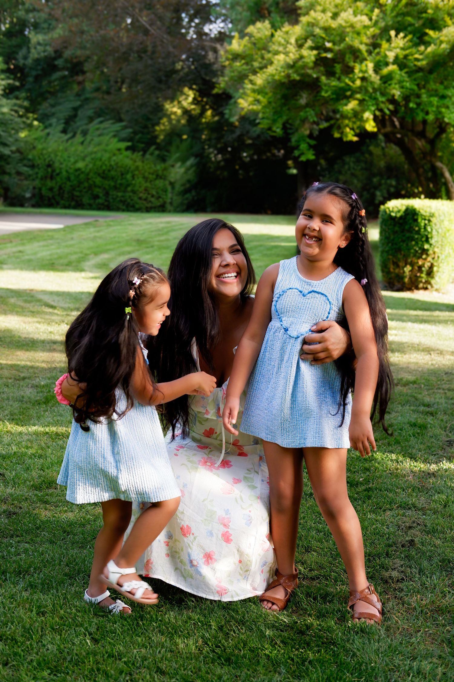 A smiling mother in a floral dress laughs with her two young daughters in light blue dresses on a green lawn.