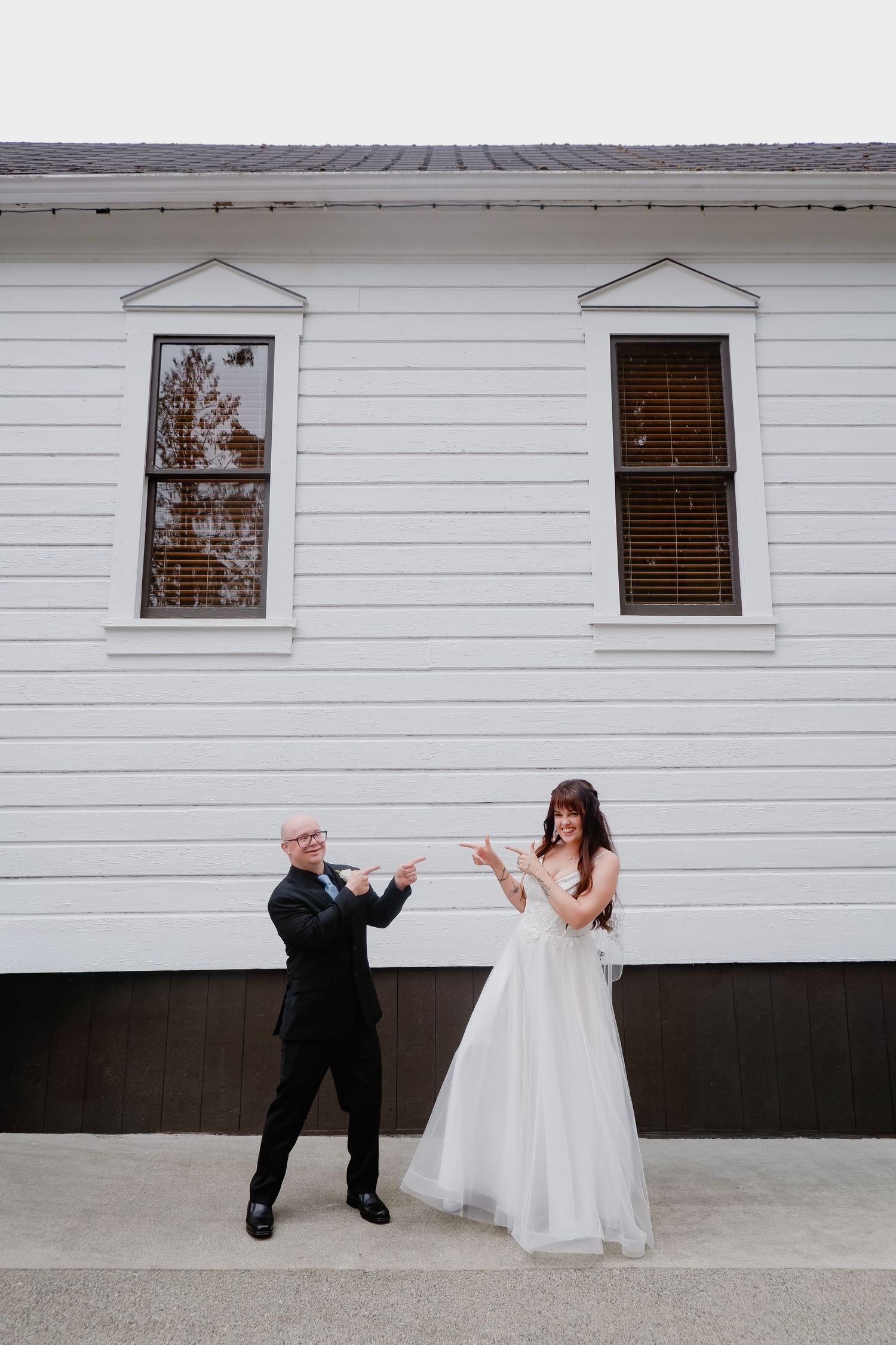 Bride and groom playfully pointing at each other in front of a white building on their wedding day.