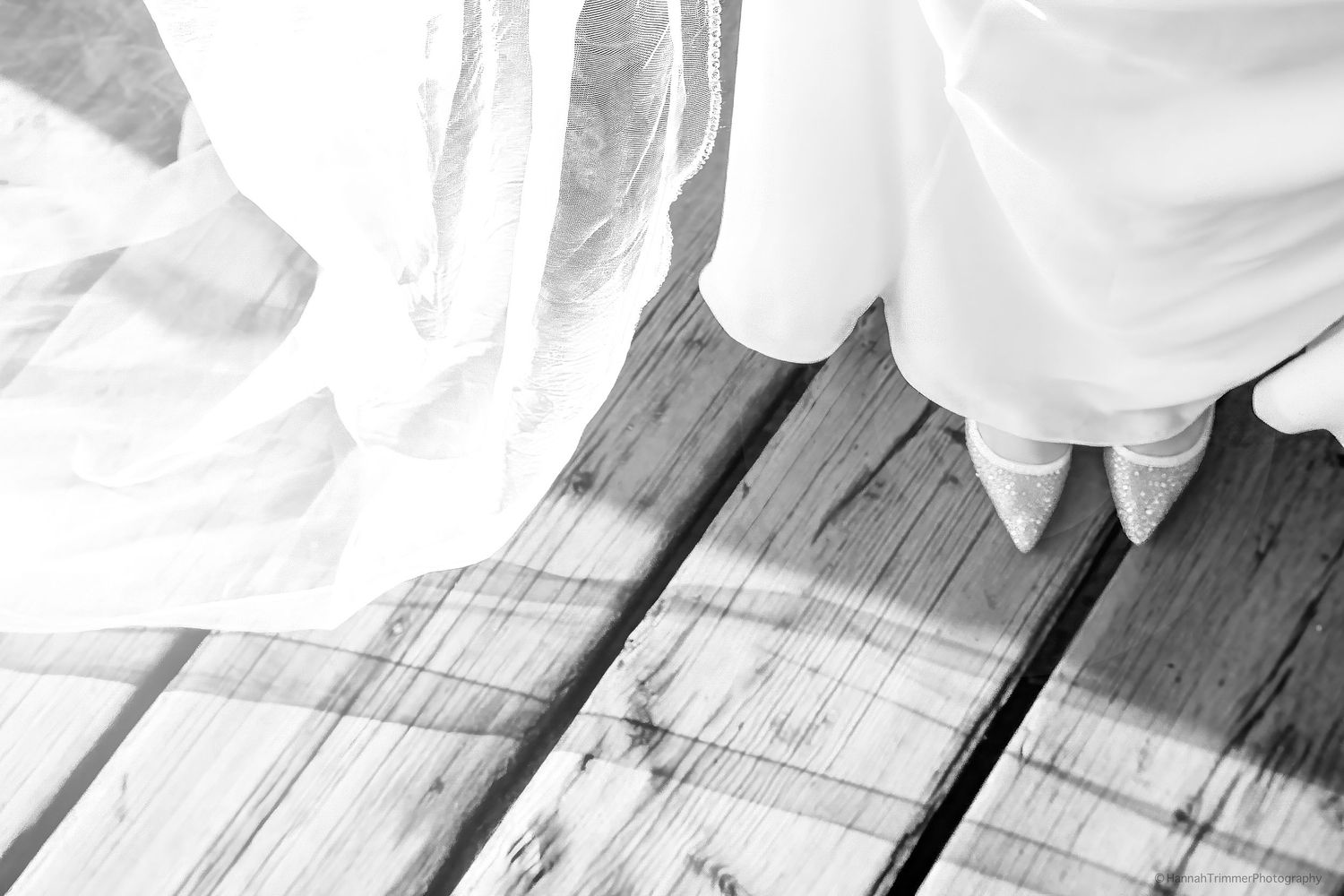 Black and white close-up of bride's white wedding dress hem on rustic wooden deck boards.
