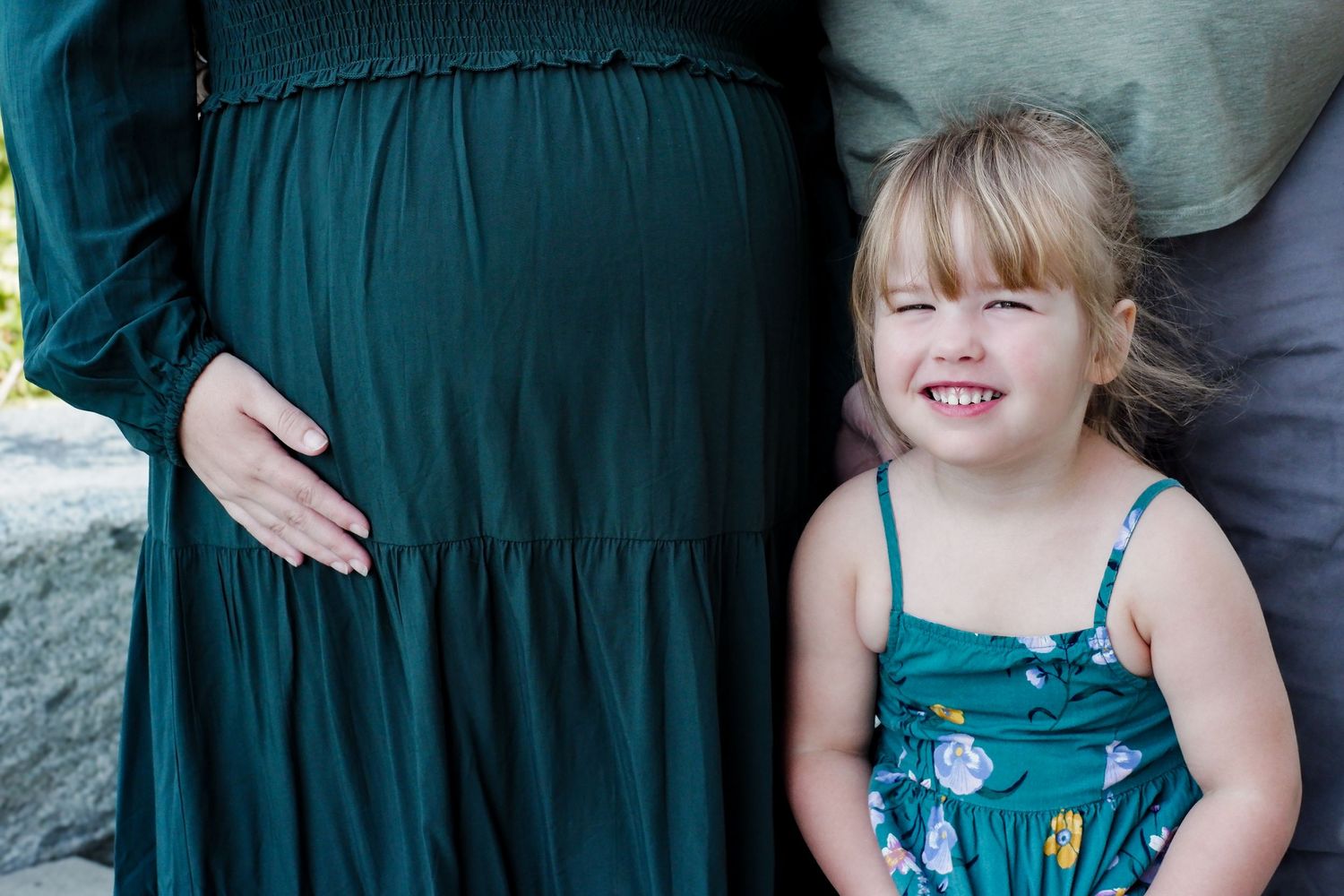 A smiling toddler girl in a teal floral dress stands beside a pregnant woman in a dark green maxi dress outdoors.