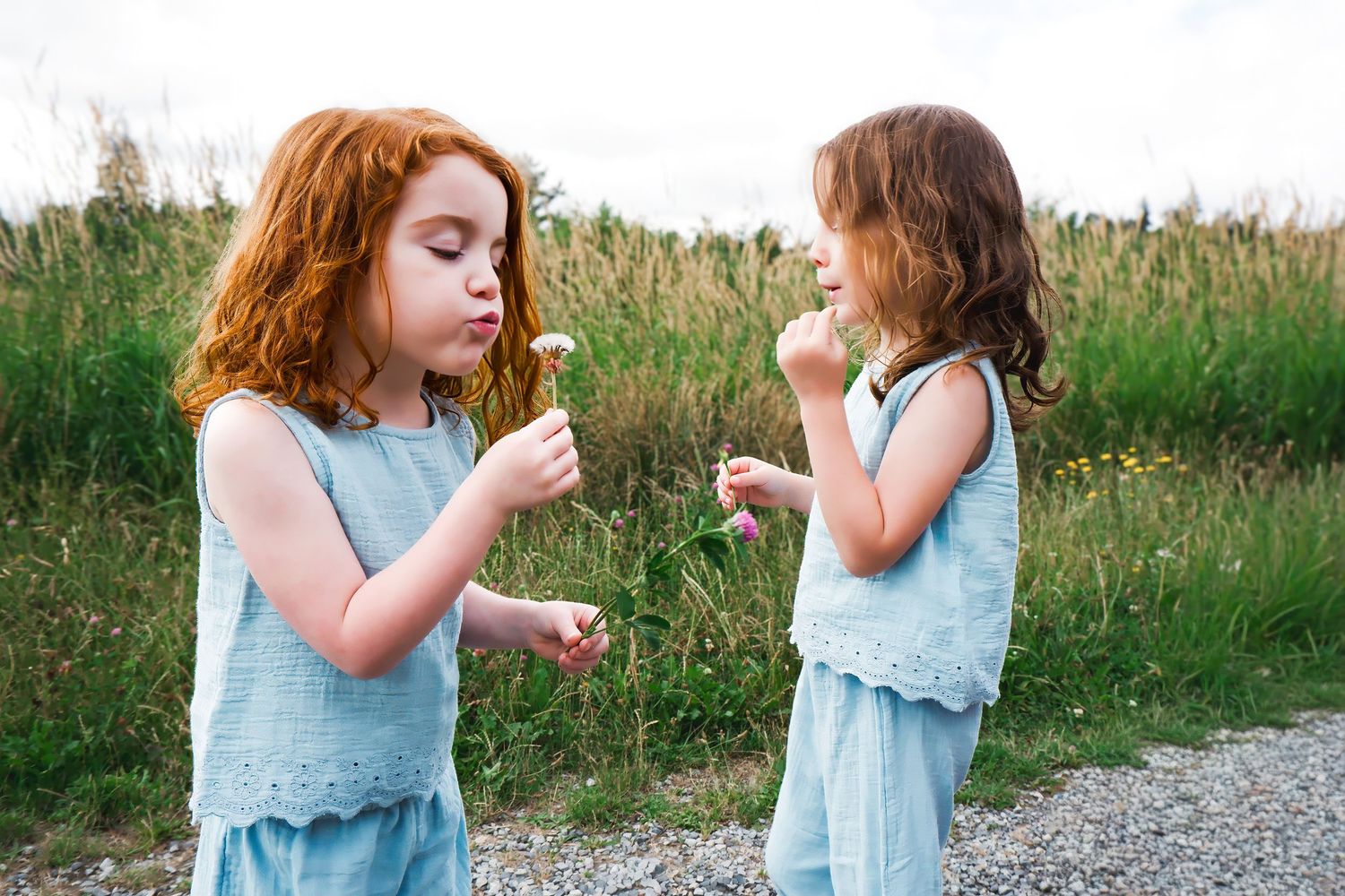 Two young girls in matching blue outfits blow dandelions together beside a grassy field on a cloudy day.