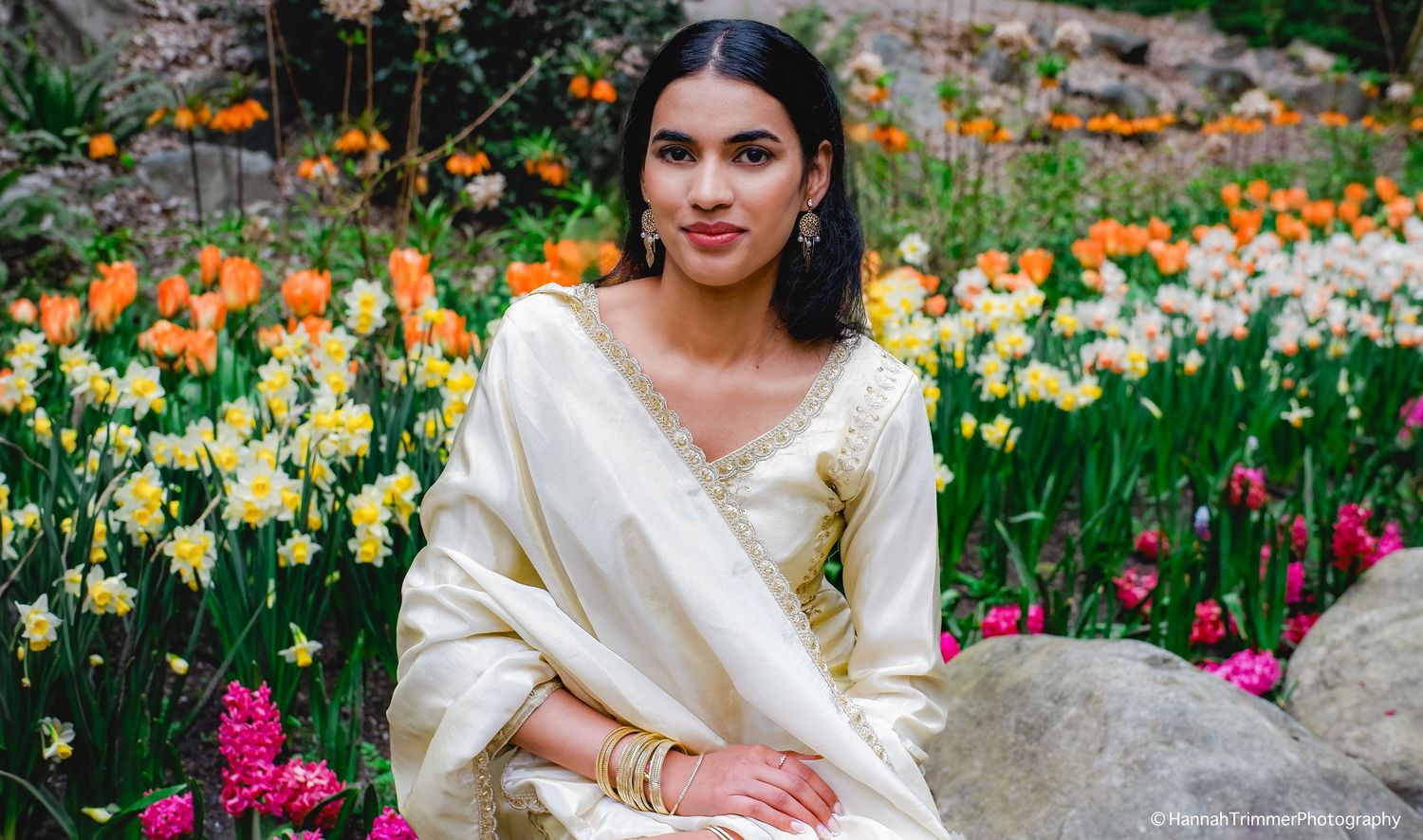 Woman in cream embroidered salwar kameez sits gracefully among colorful spring garden flowers.