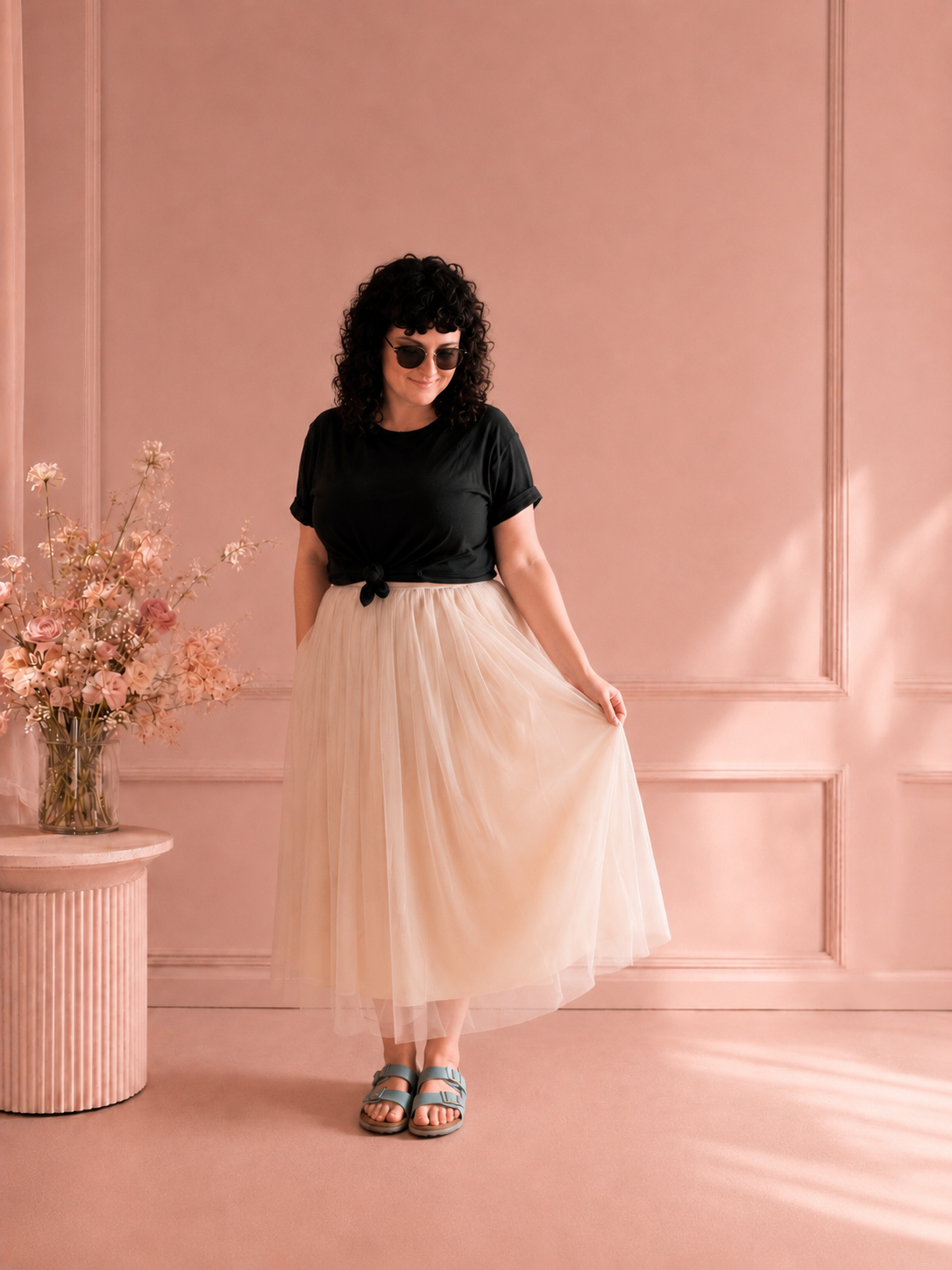 Woman in black top and cream tulle skirt poses in pink-walled studio.