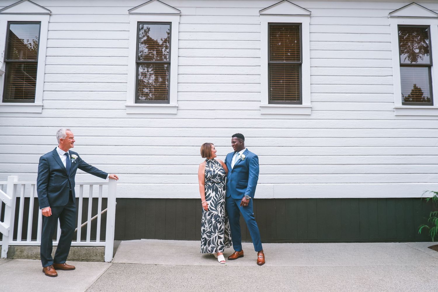Three people in formal attire stand outside a white building, with two men in suits and a woman in a floral dress.