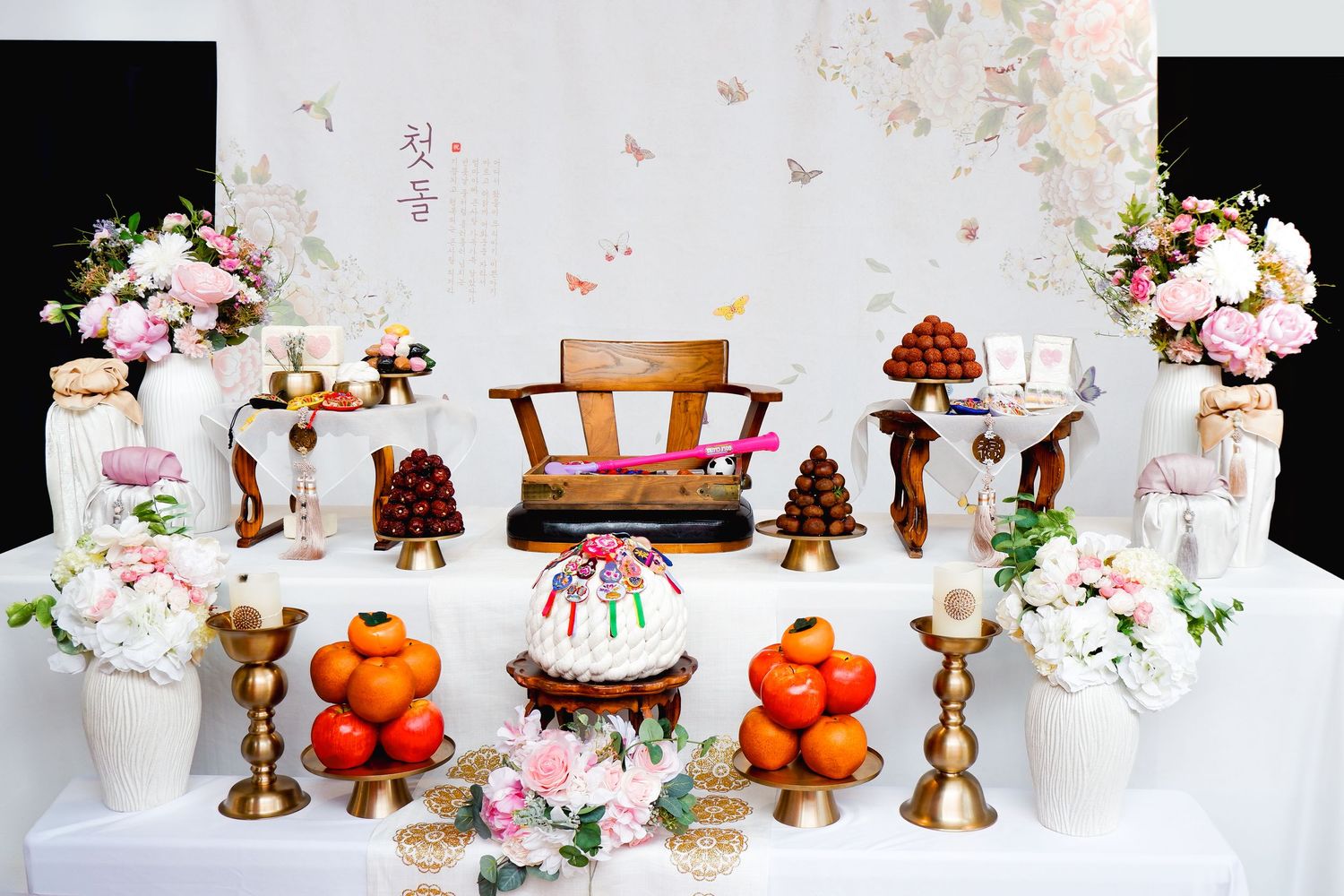 Traditional Korean wedding table display with floral arrangements, brass candlesticks, persimmons, and ornate decorations on white cloth.