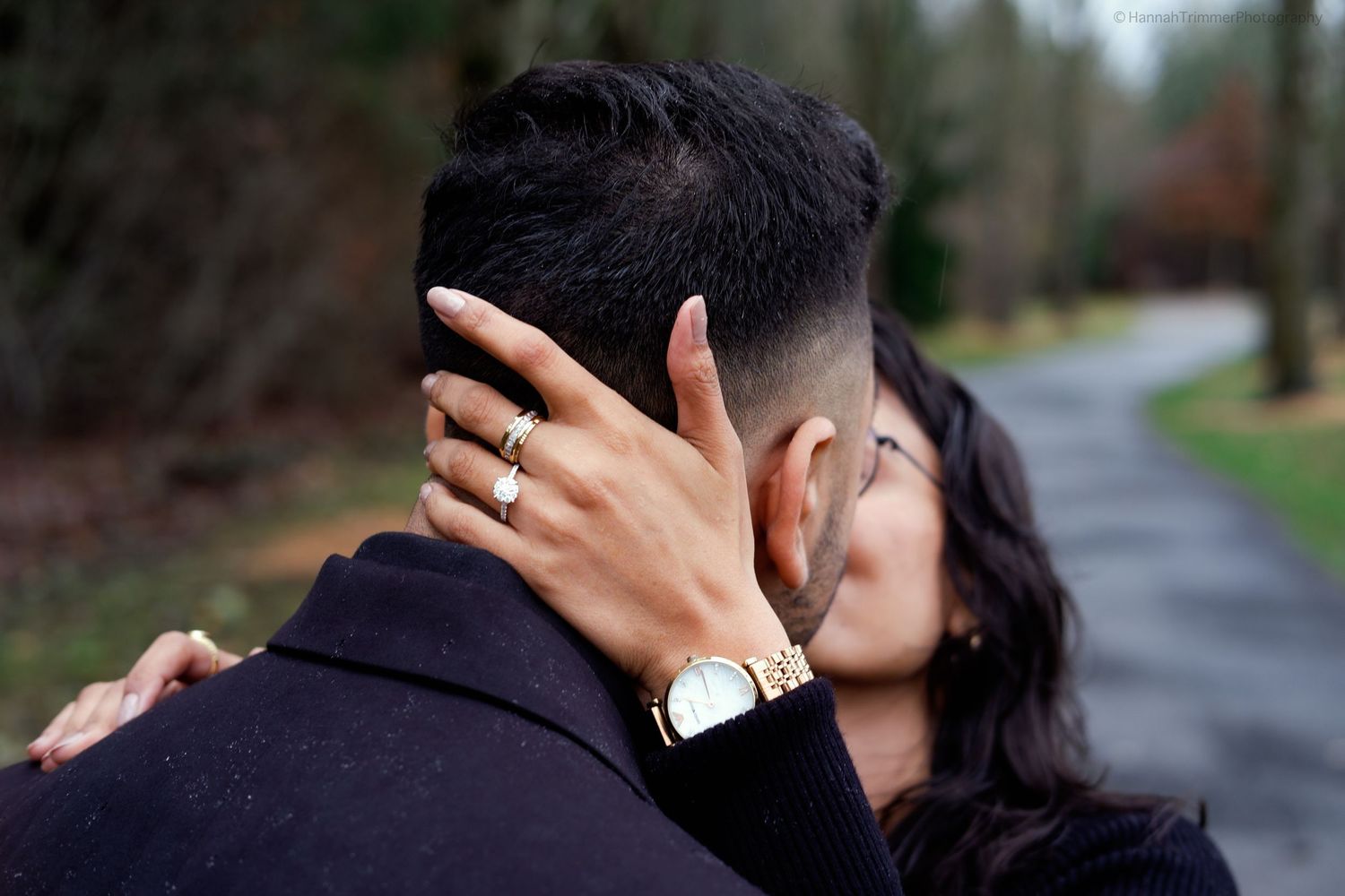 Close-up of newly engaged couple kissing, showcasing engagement ring and gold watch outdoors.