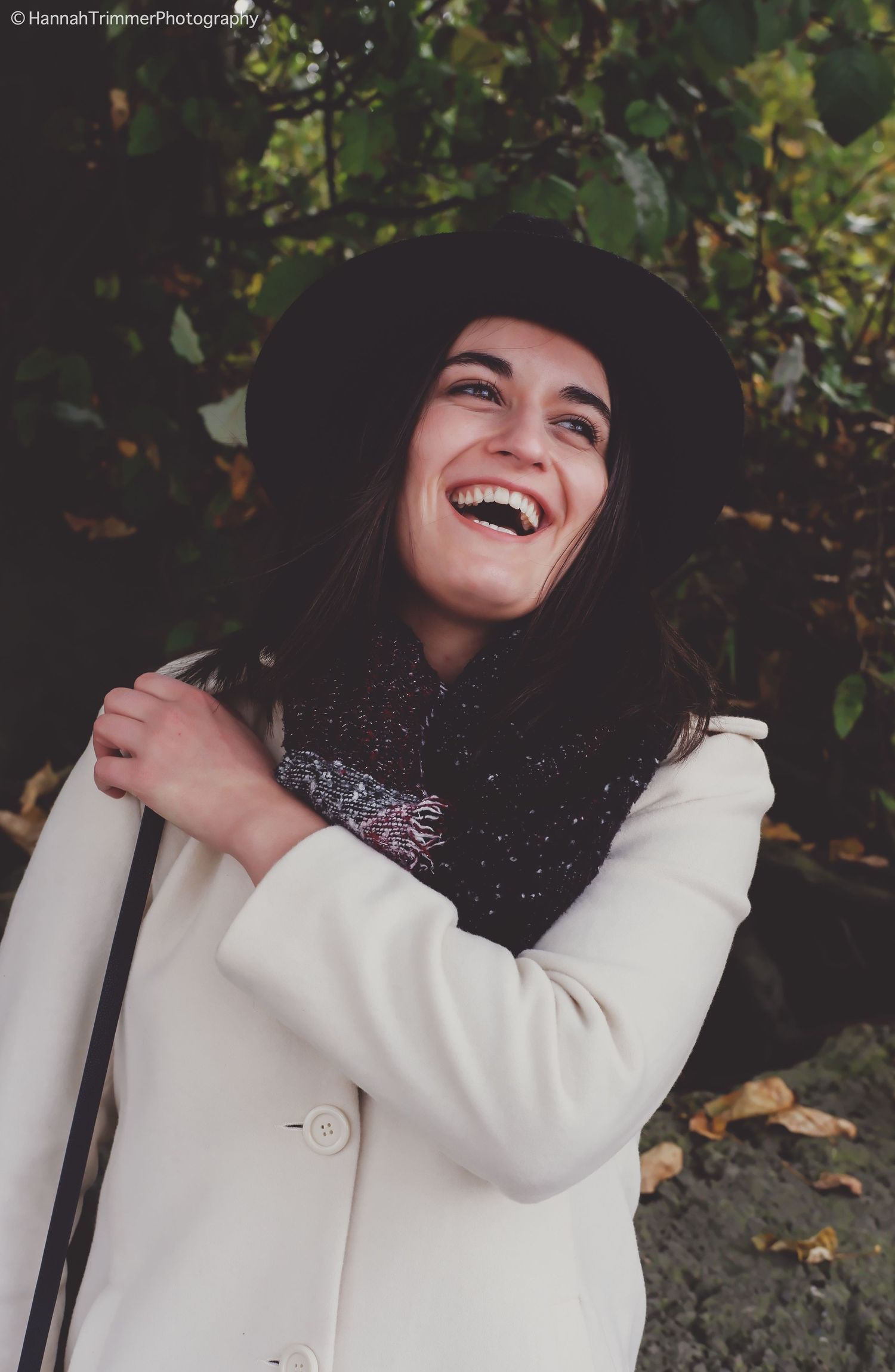 Joyful woman in white coat and black hat laughing outdoors among autumn foliage.