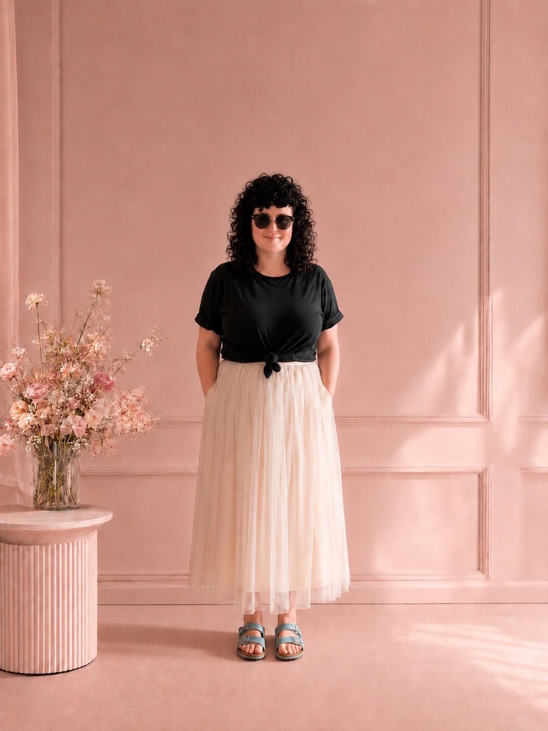 Curly-haired woman in black top and cream tulle skirt posing in pink studio.