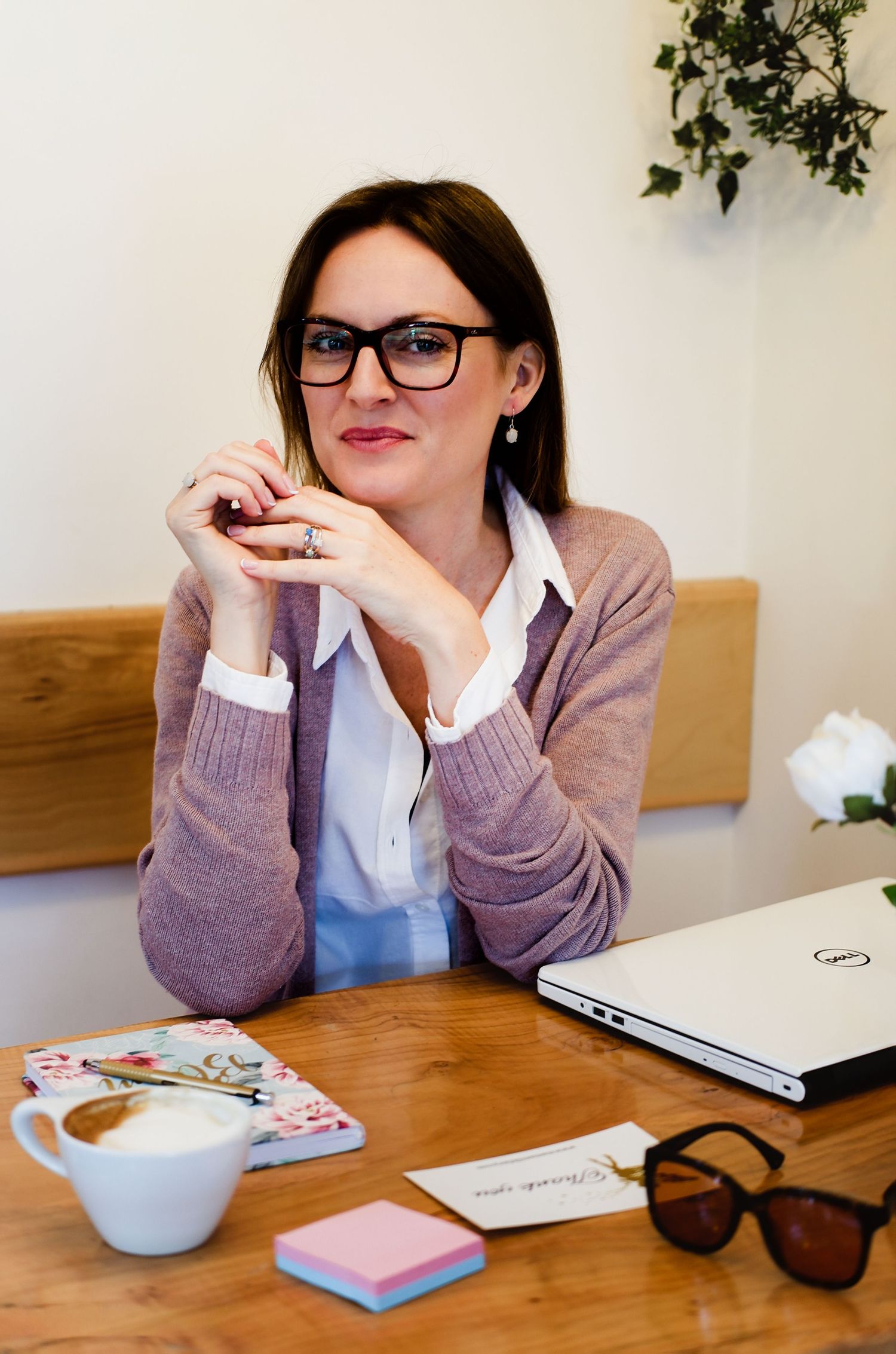 Professional woman with glasses sits at wooden desk with laptop and coffee in bright cafe.