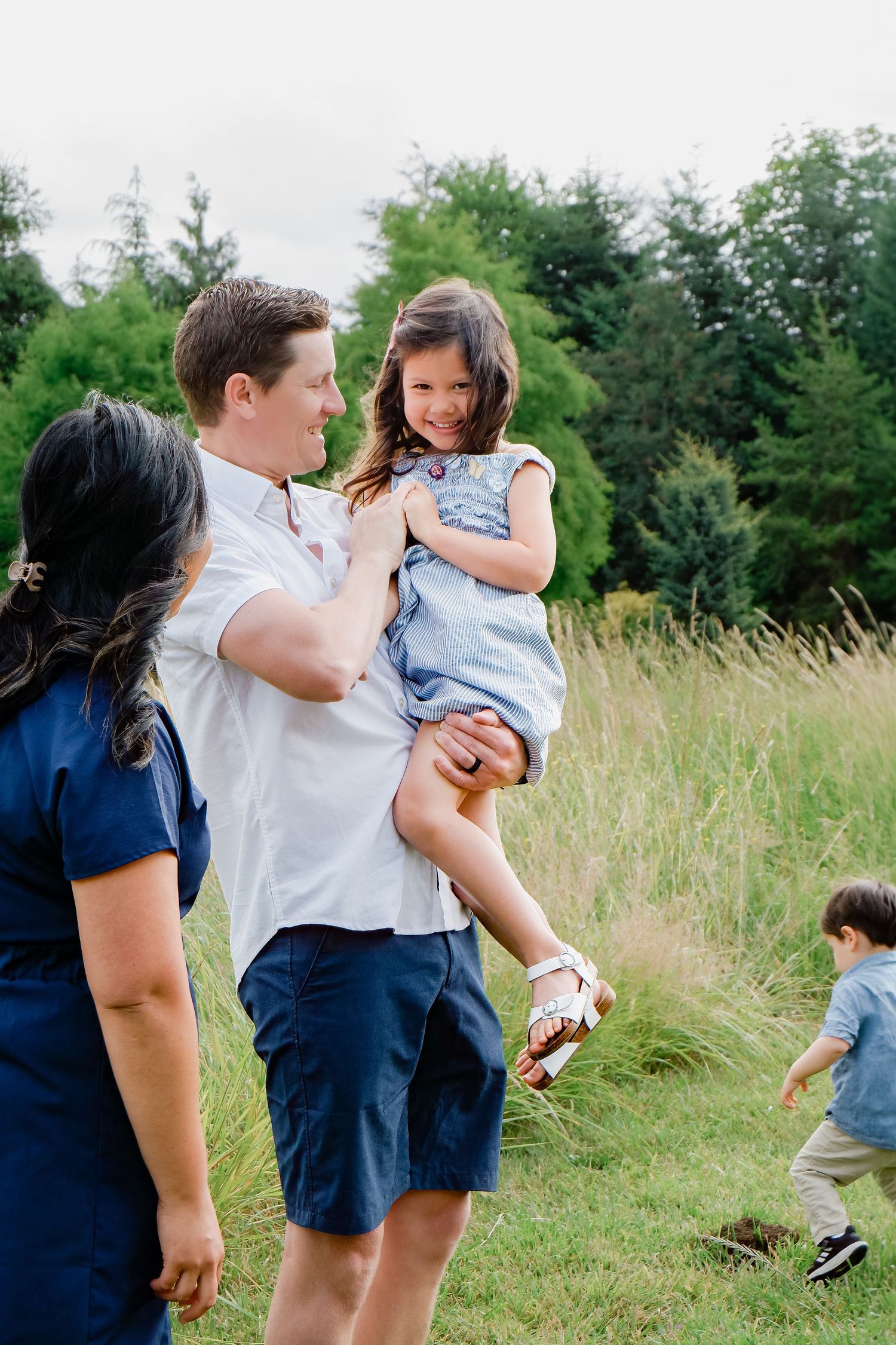 A father lifts his laughing daughter in a blue dress while mother and toddler son enjoy a lush meadow.