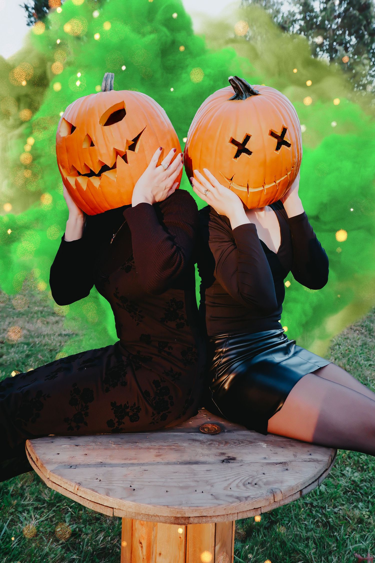 Two women in black dresses hold carved Halloween pumpkins over their faces outdoors with green smoke and bokeh lights.