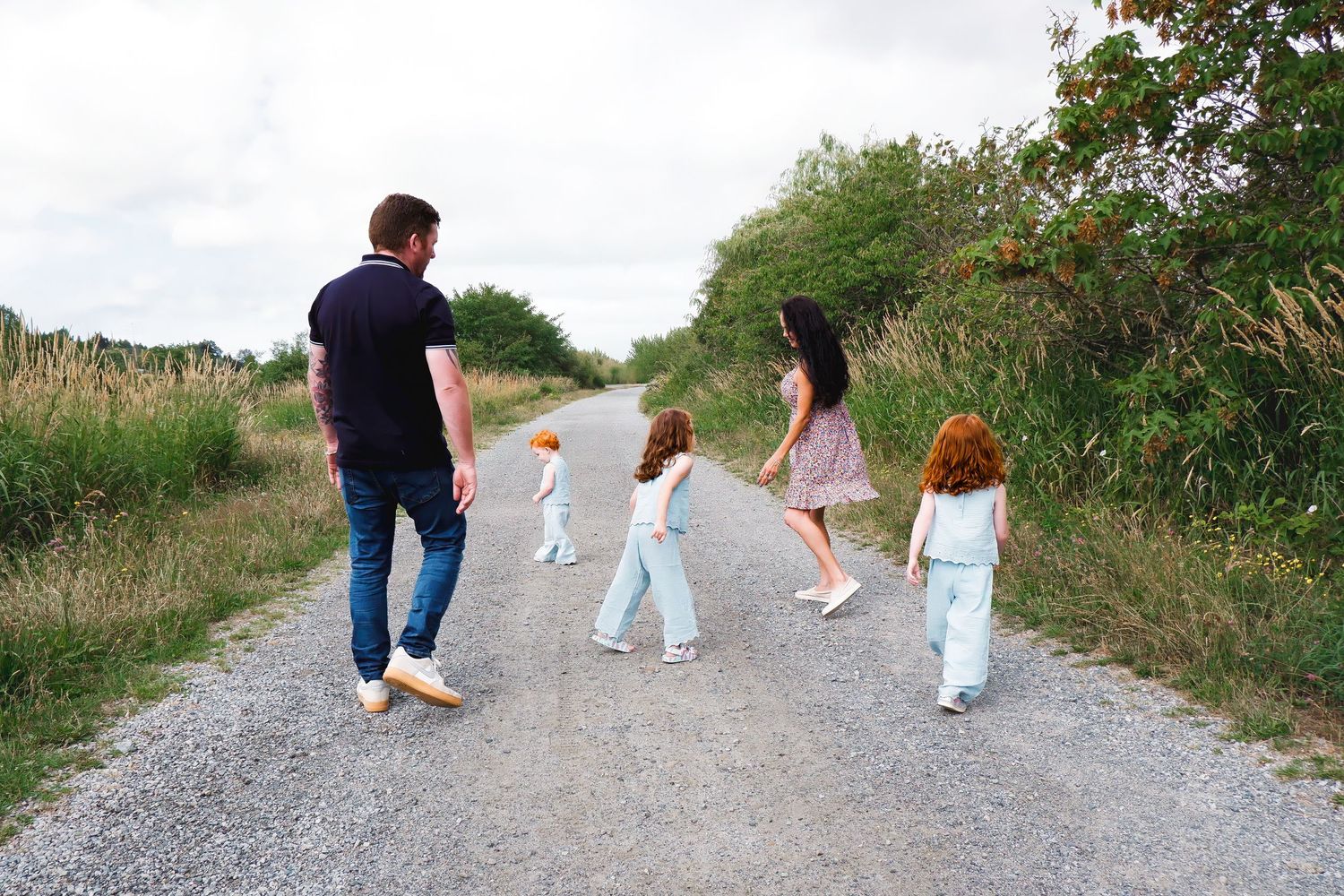 Family of five walking away on a rural gravel path surrounded by lush green vegetation.
