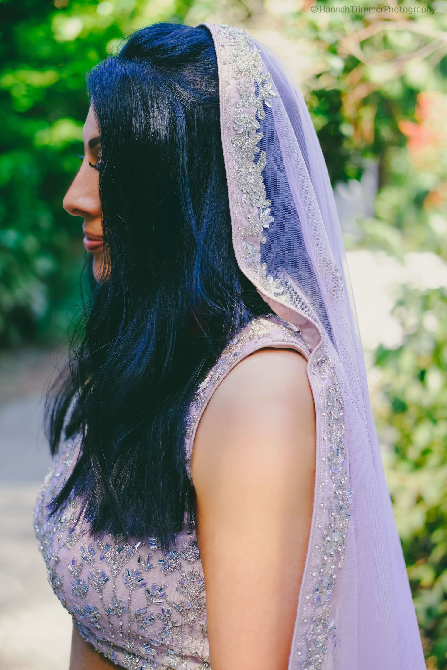 Bride in embellished lavender lehenga with matching veil shows elegant hair in garden setting.