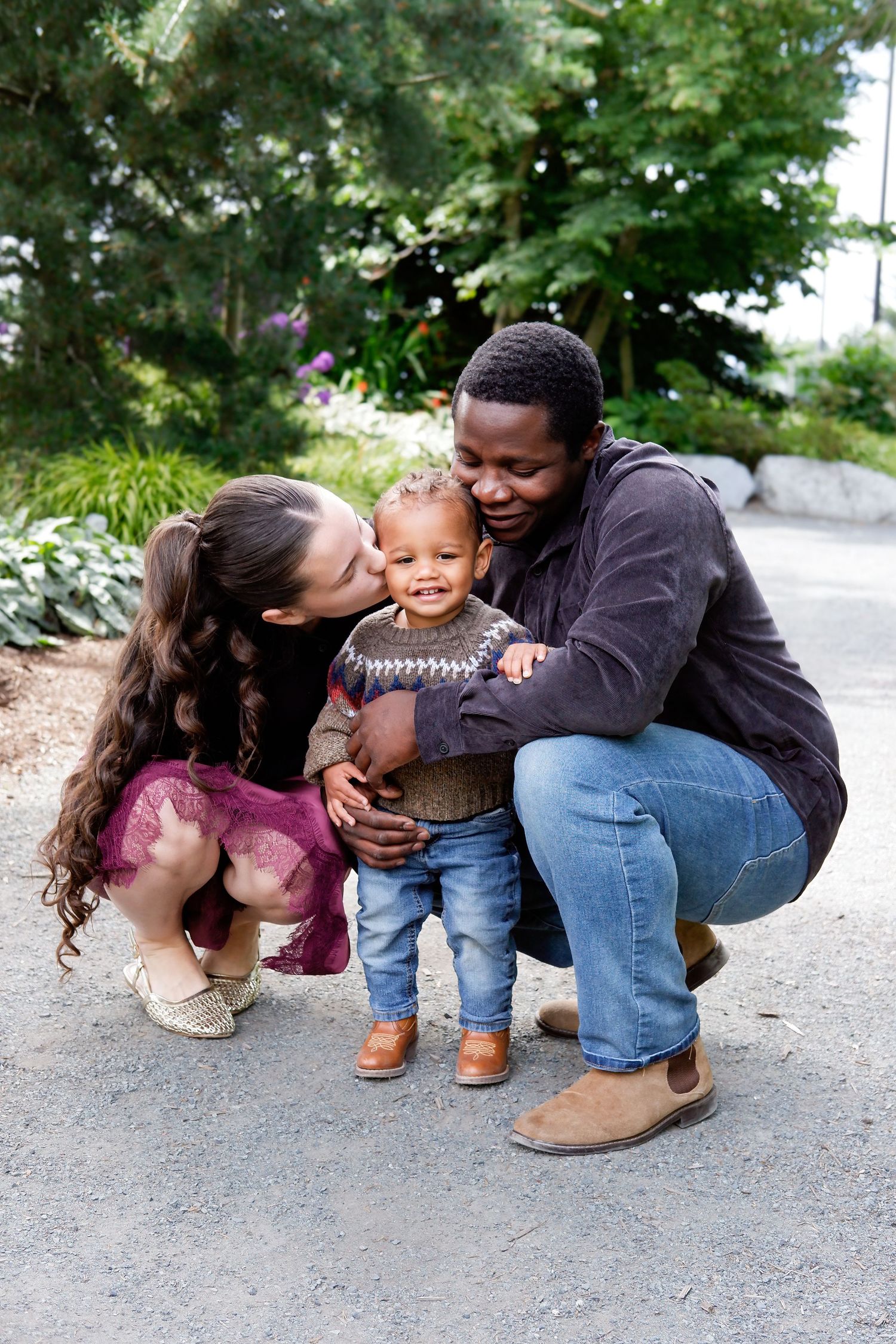 Interracial parents crouching to hug and kiss their happy toddler on a garden pathway.