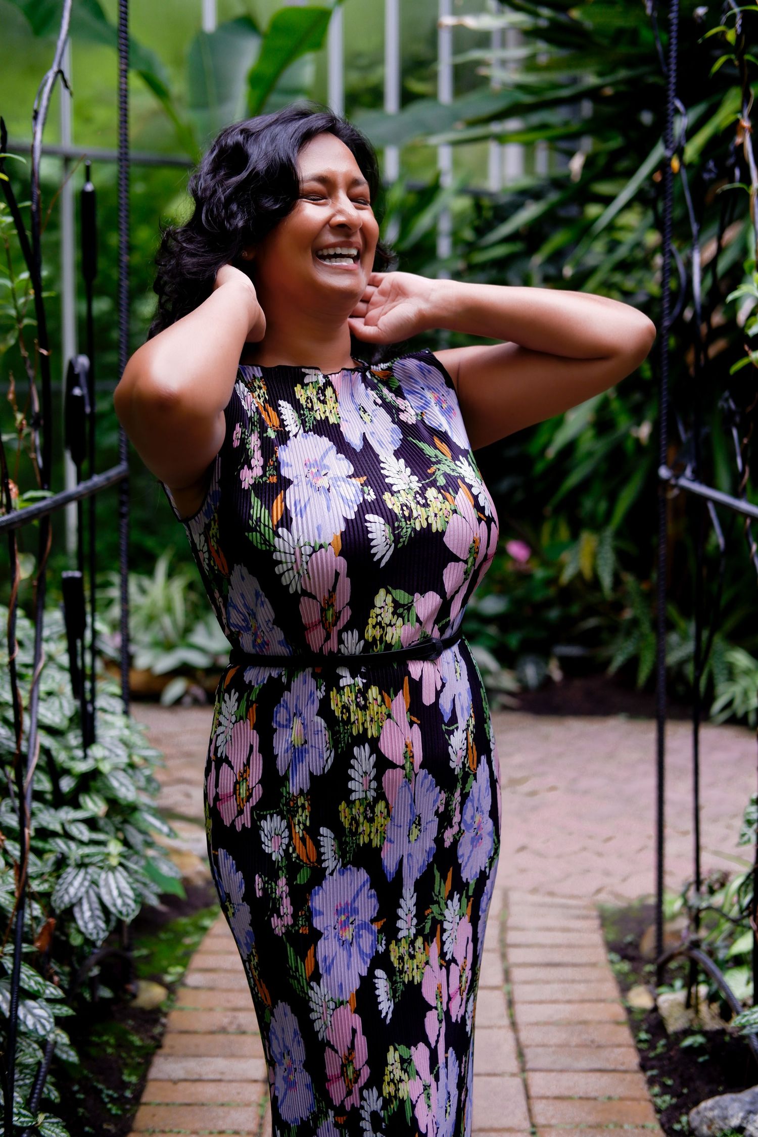 Woman in black floral fitted dress laughing joyfully inside a lush greenhouse garden.