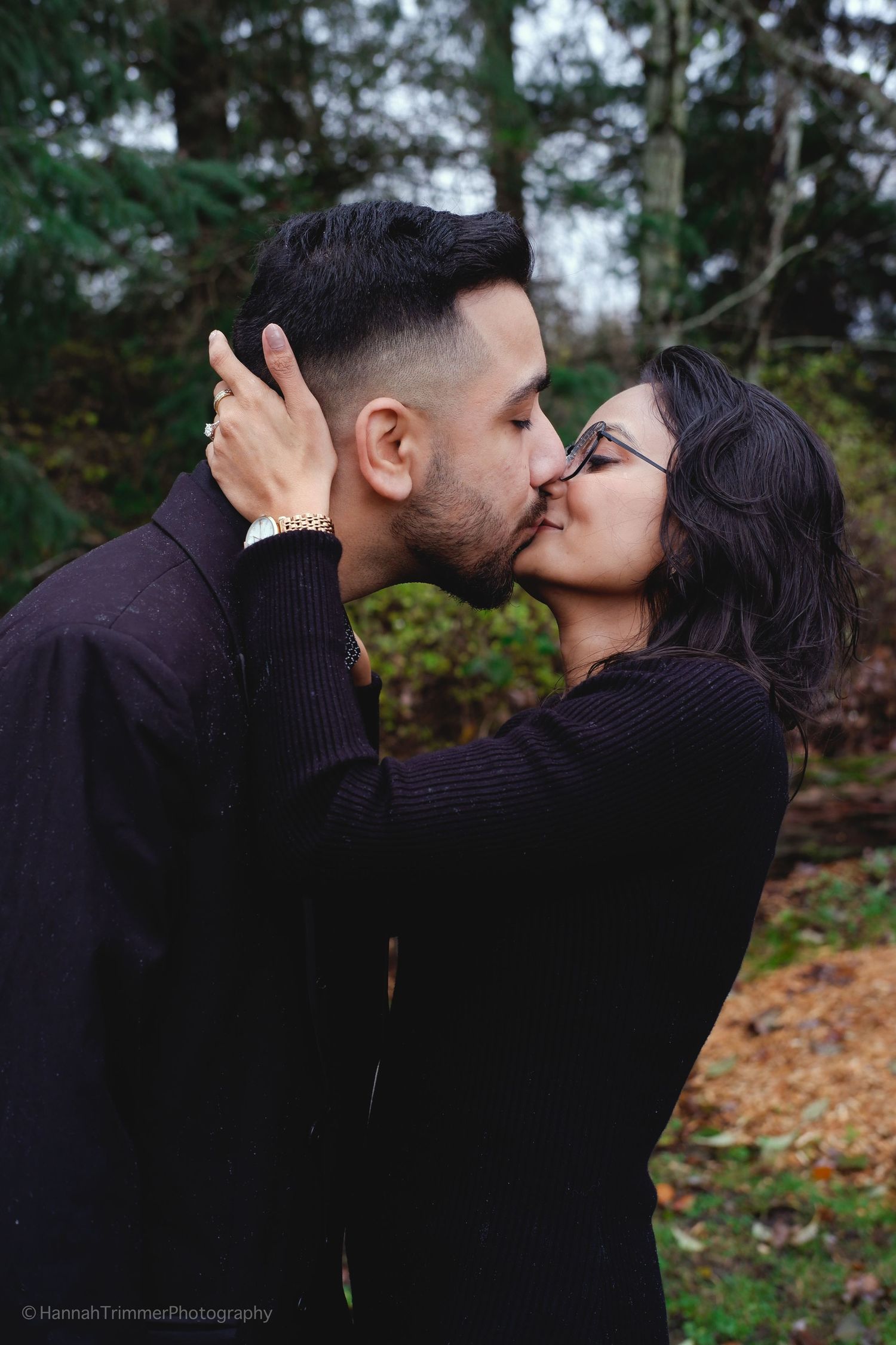 Couple sharing a passionate kiss outdoors in a forest setting wearing matching black outfits.