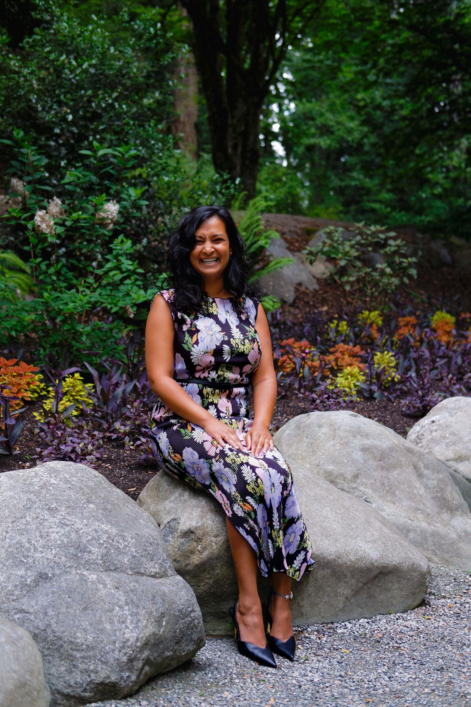 Smiling woman in floral dress sits on large rocks surrounded by lush garden greenery and colorful flowers.