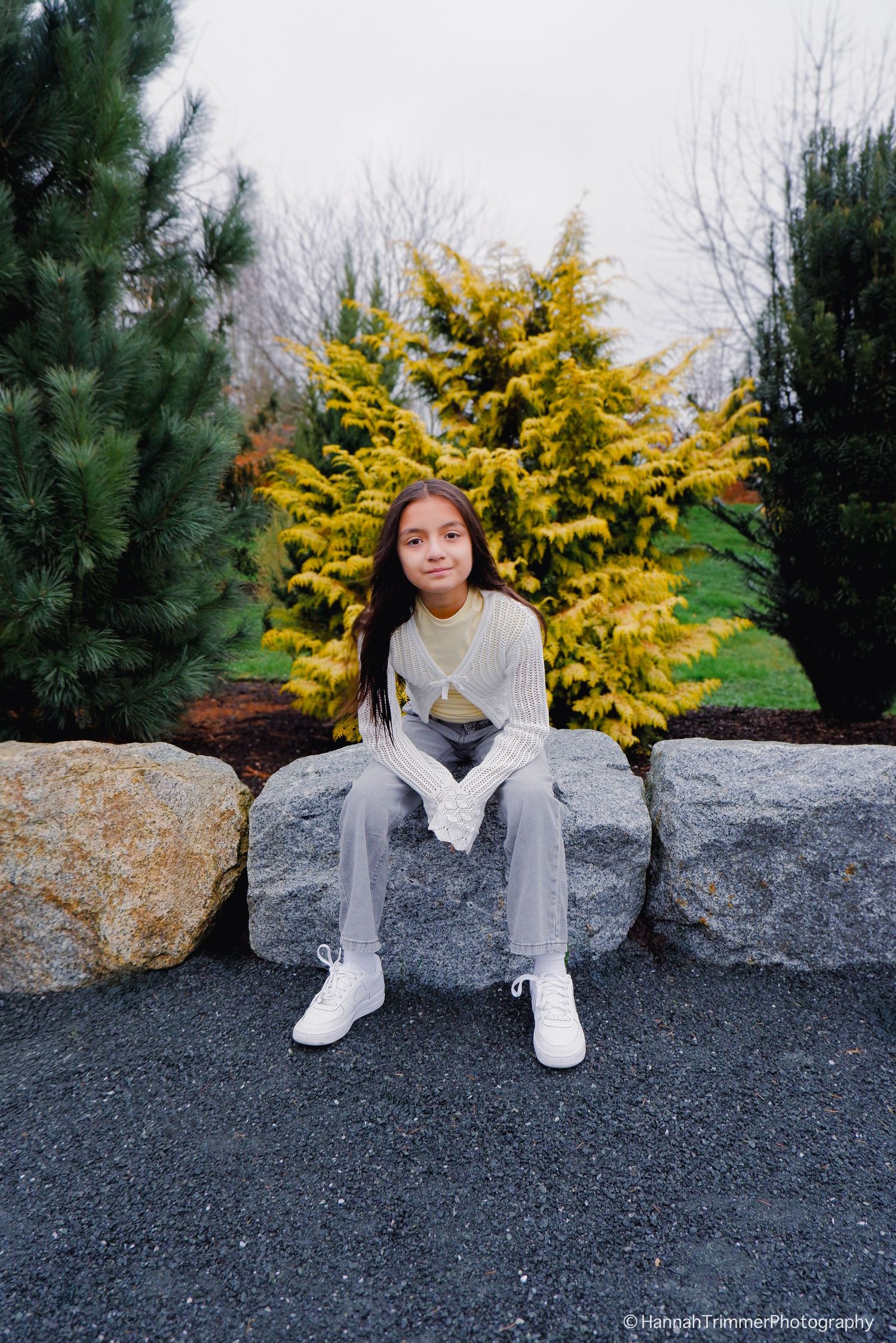 Young girl in white top and gray jeans sitting on rocks near golden evergreen trees.