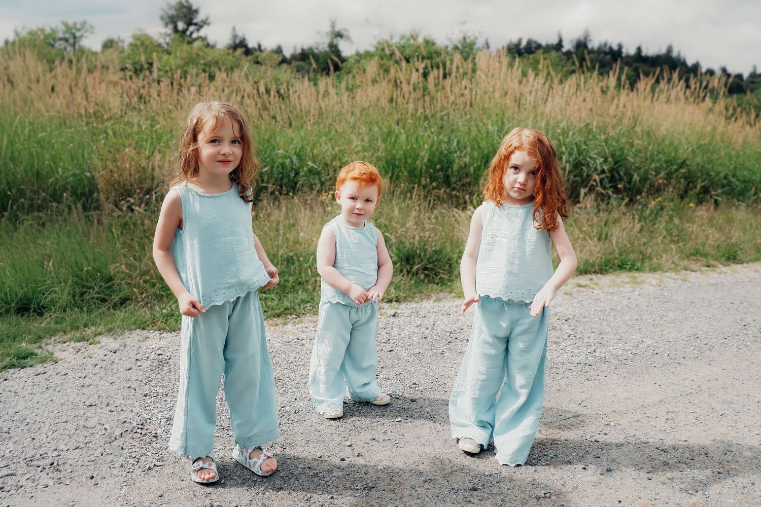 Three young girls in matching light blue outfits standing on a gravel path surrounded by green fields.