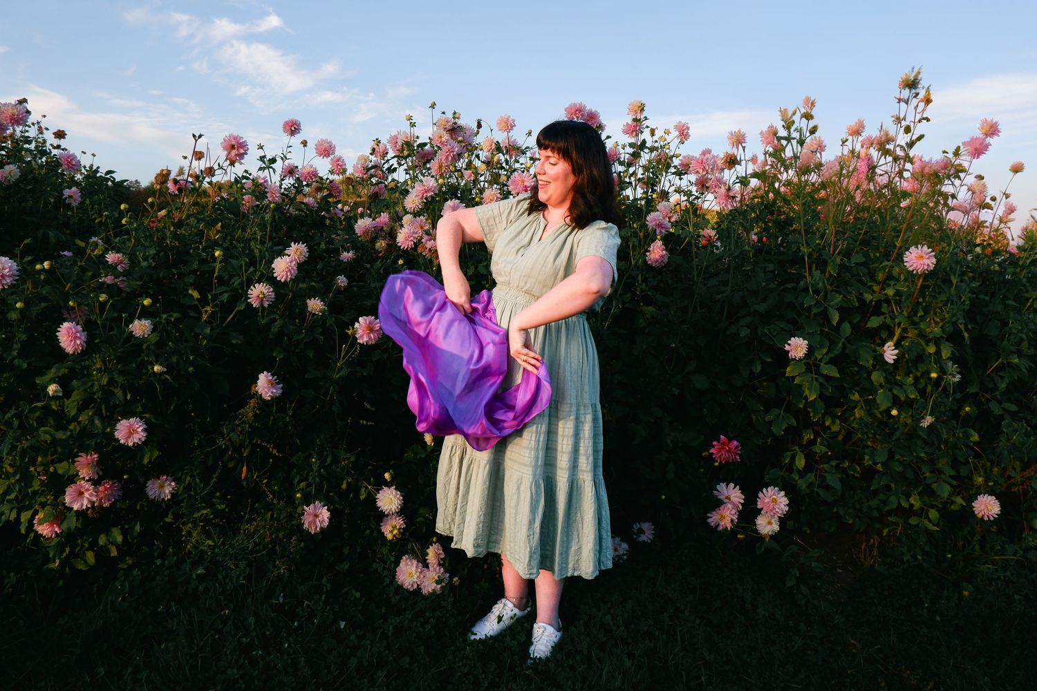 Woman in green dress holding purple balloons among blooming pink roses in a garden at golden hour.