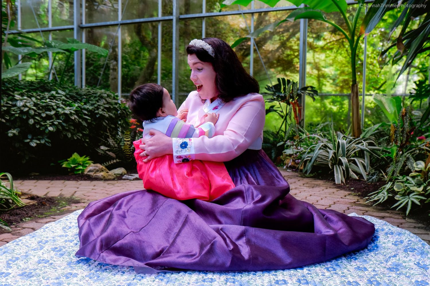 Woman in traditional Korean hanbok holds child in greenhouse setting.
