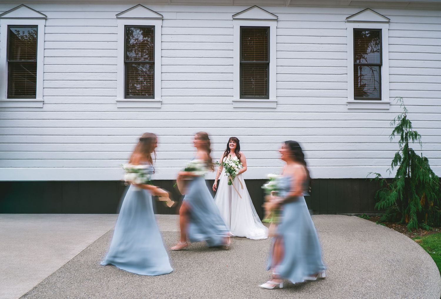Bride in white gown poses with blurred bridesmaids in blue dresses outside a white building.