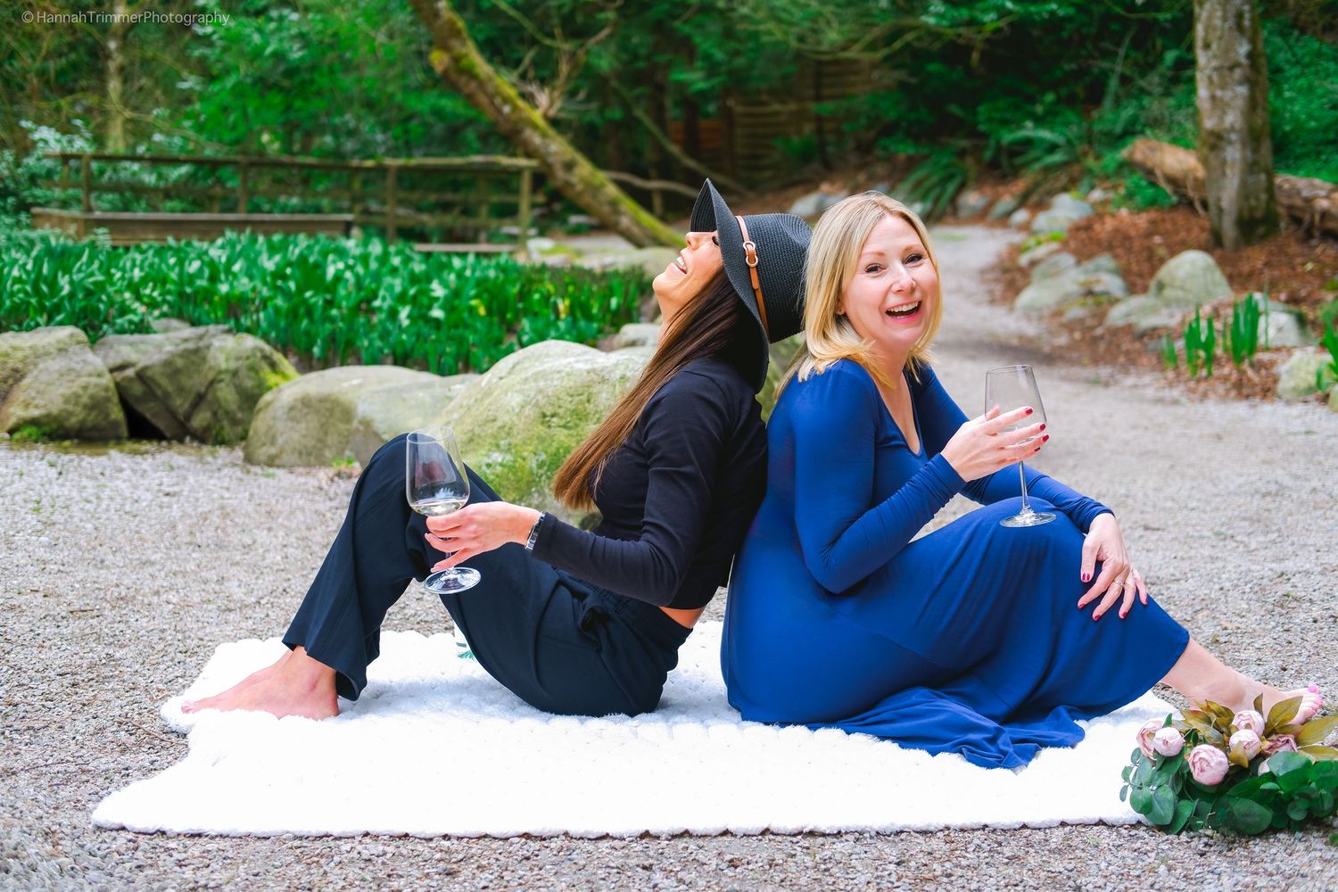 Two women sitting back to back on a white blanket outdoors holding wine glasses and laughing.