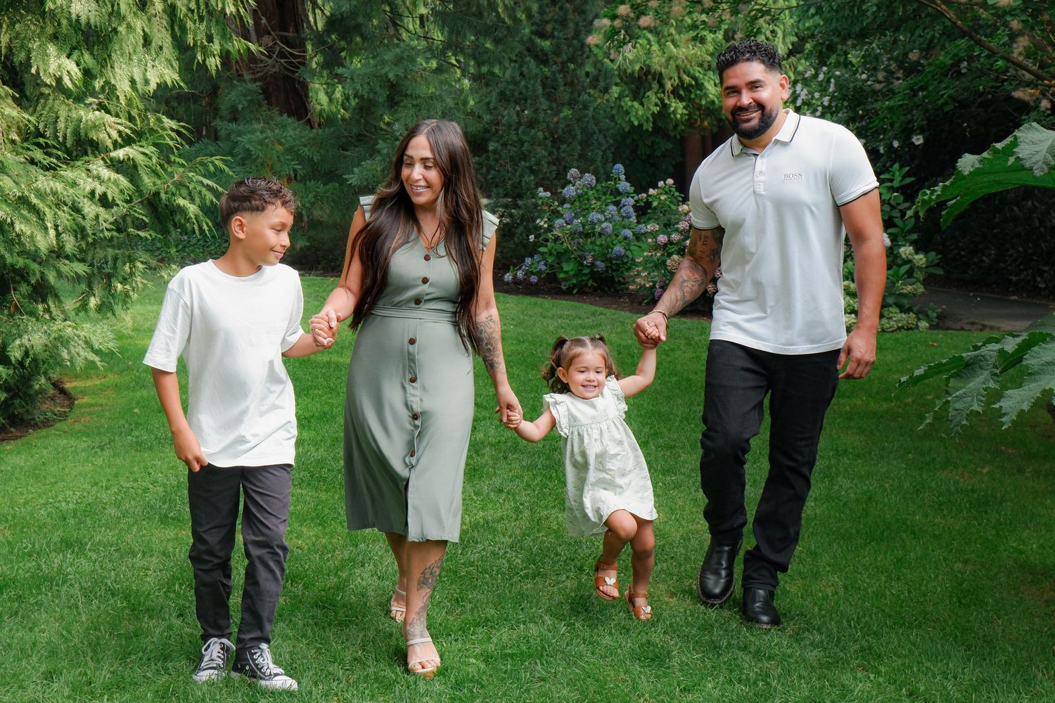 Happy family of four walking hand-in-hand on lush green lawn surrounded by trees in a sunny garden.