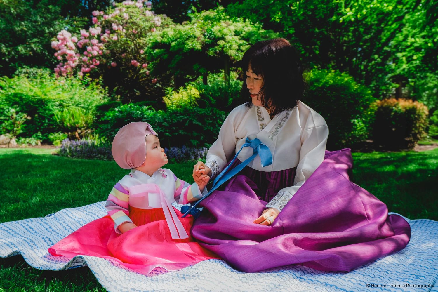 Mother and baby in traditional Korean hanbok sitting on blanket in lush green garden.