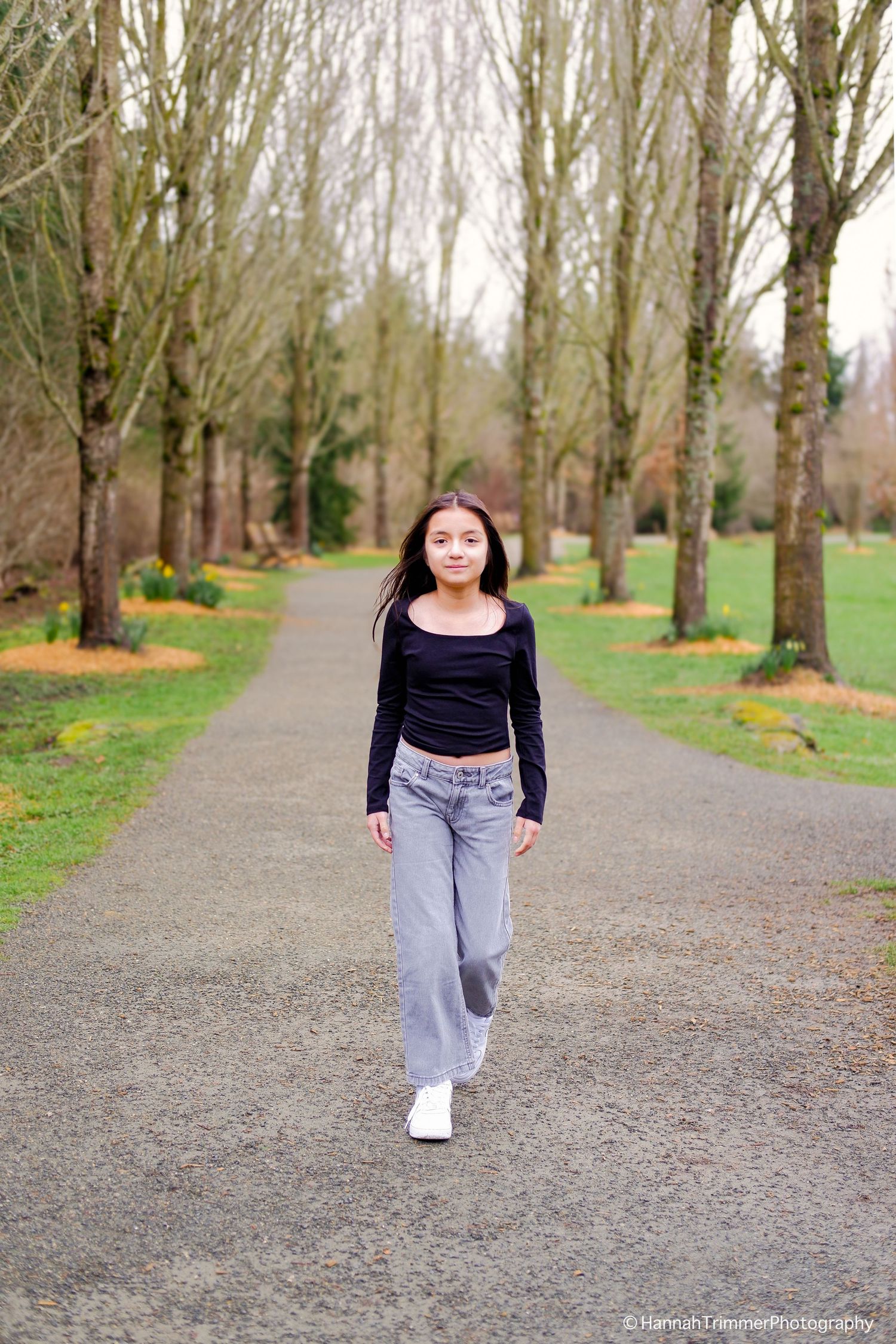 Teen girl in black top and gray jeans walking along tree-lined park pathway.