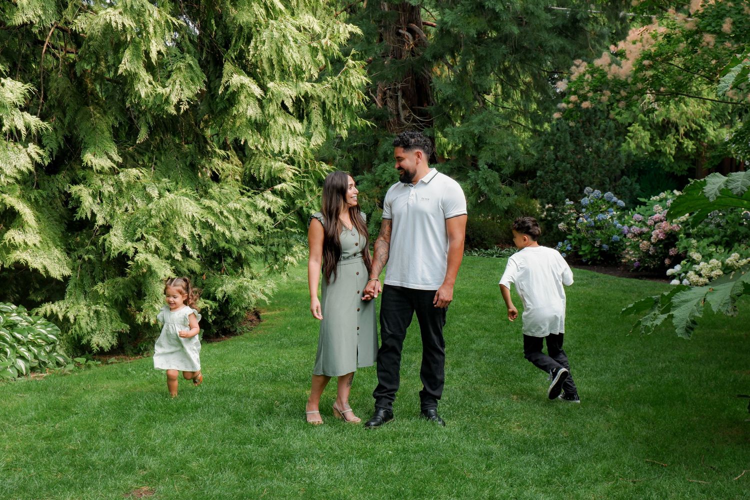 A family of four enjoys a lush green garden, with parents holding hands while two young children play around them.