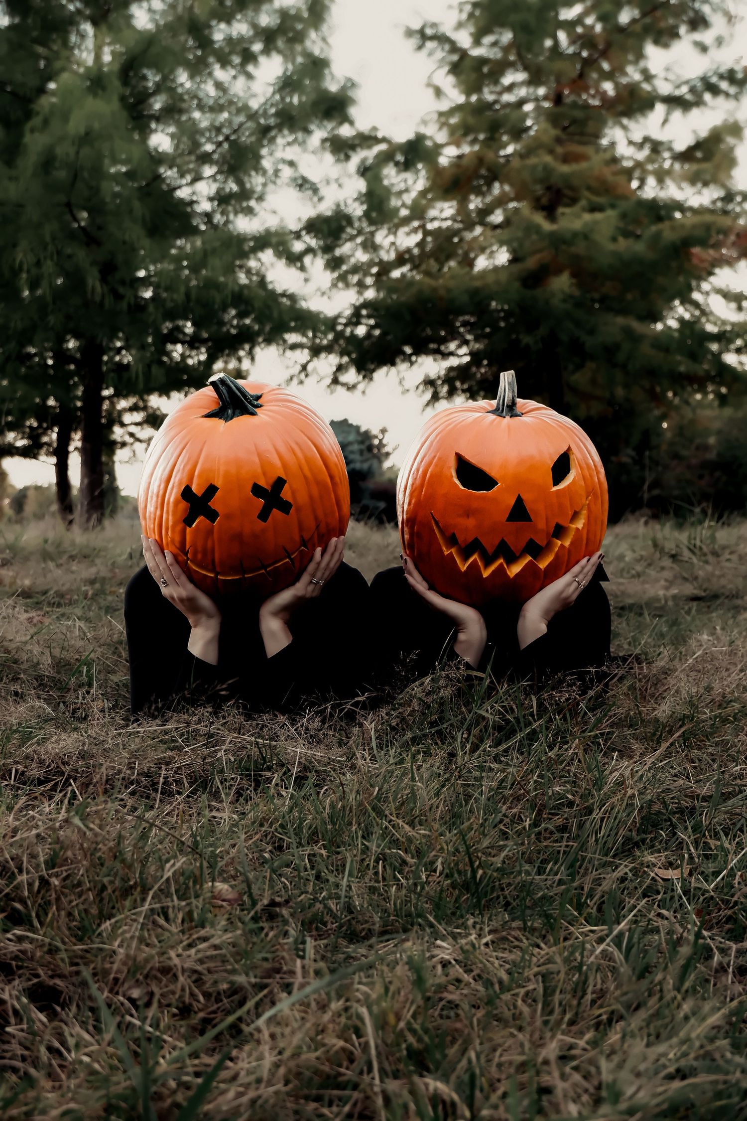 Two people holding carved Halloween jack-o-lantern pumpkins over their faces outdoors in a spooky autumn setting.