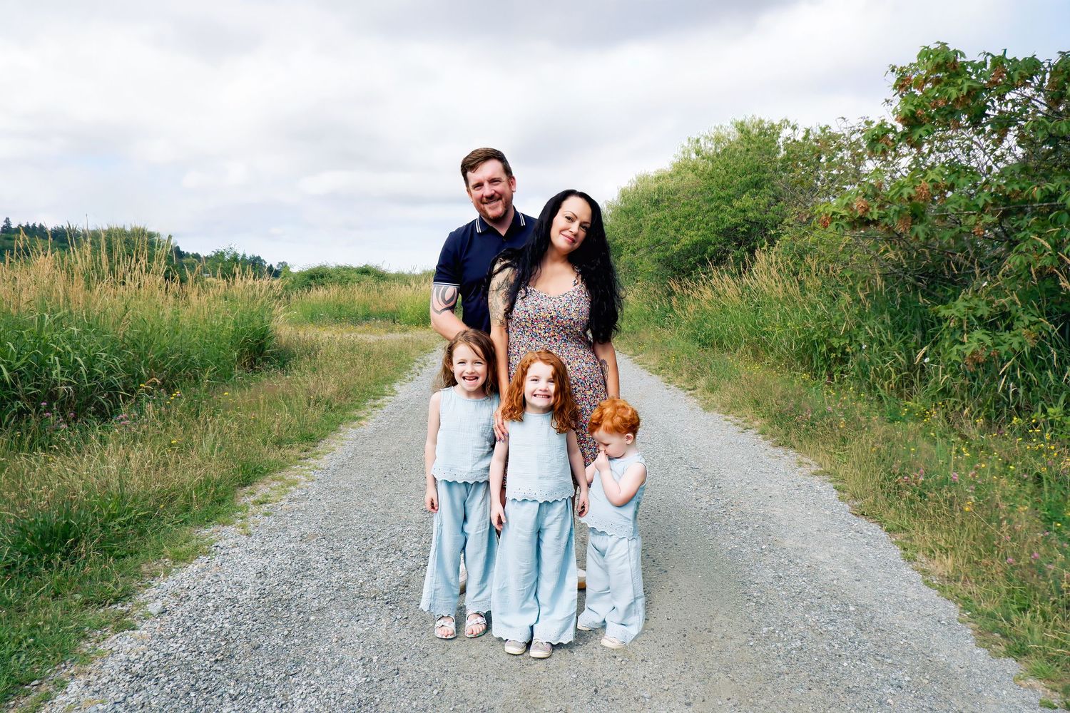 Happy family of five posing together on a gravel path with green fields behind them.