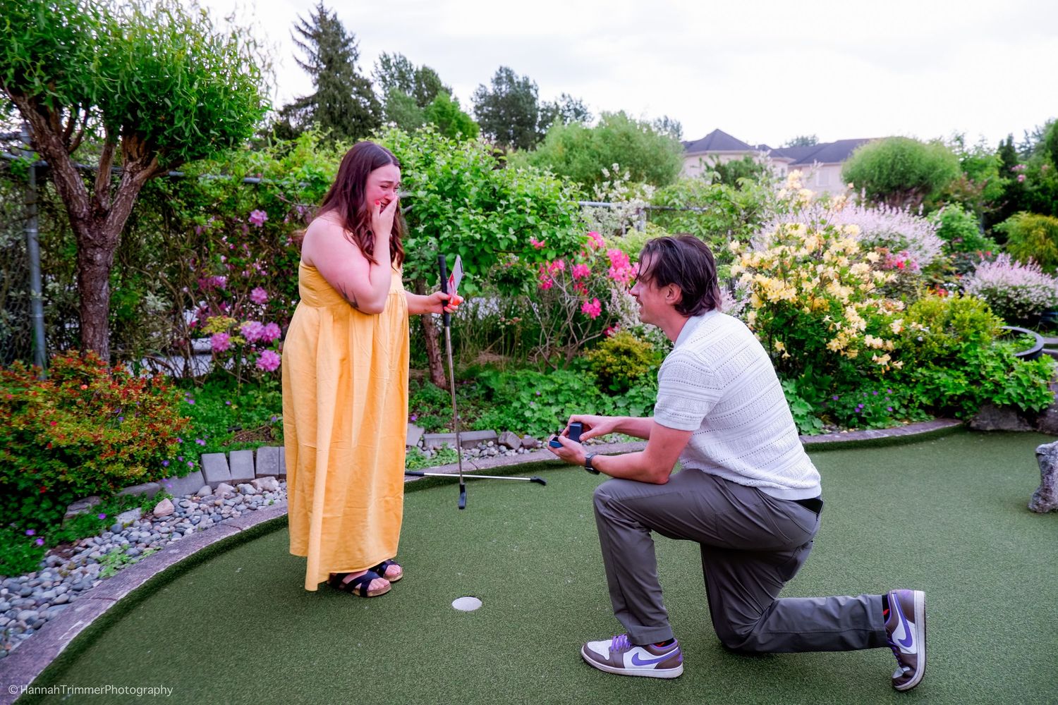 Man kneels to propose to surprised woman in yellow dress on mini golf course surrounded by flowers.
