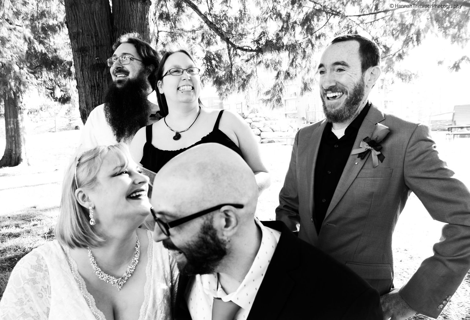 Black and white photo of laughing bride and bearded groom with two friends celebrating under a tree.
