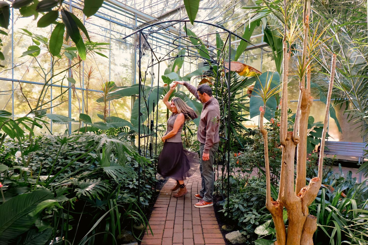 Couple exploring a lush tropical greenhouse with brick pathway, surrounded by exotic plants and glass ceiling.