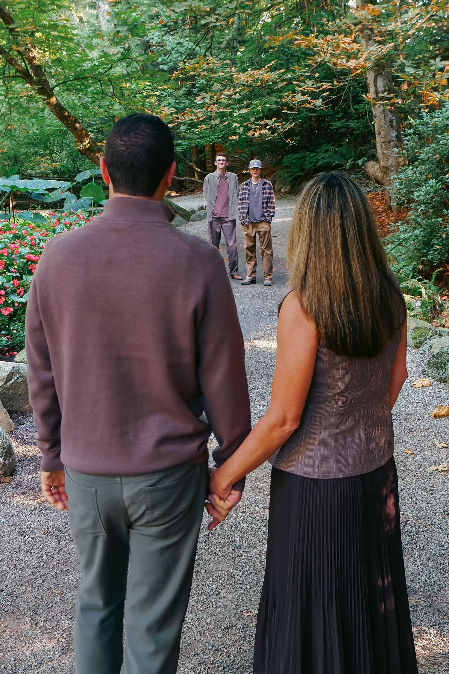 A couple holds hands on a garden path facing two young men standing among lush green trees and red flowers.