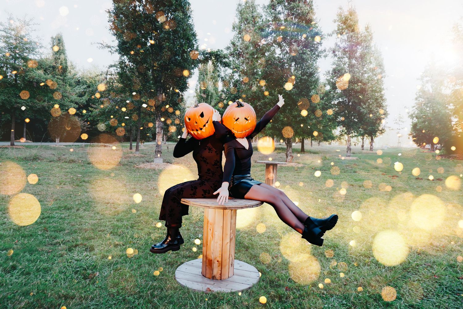 Two people wearing Halloween pumpkin heads sit on a wooden table outdoors with bokeh lights in a park.